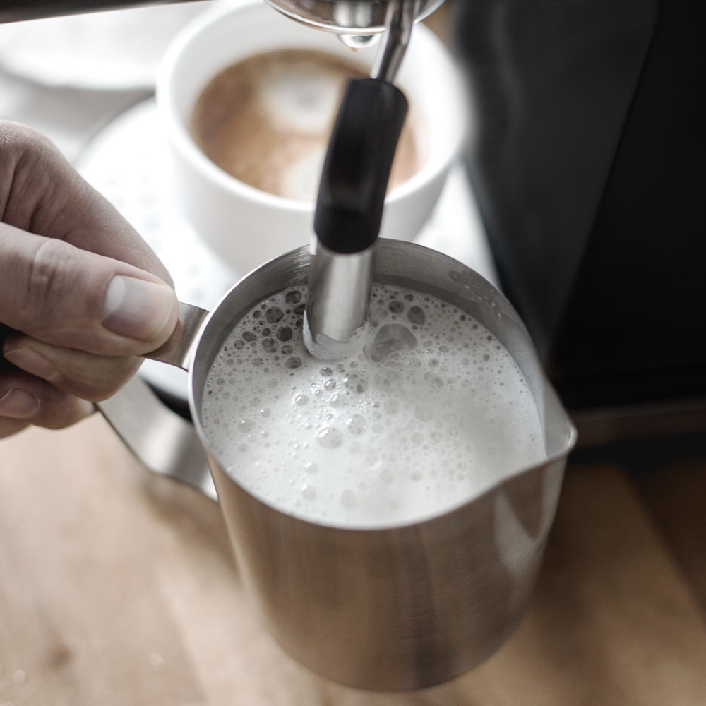 Barista spienia mleko do srebrnego dzbanka, z filiżanką kawy w tle.