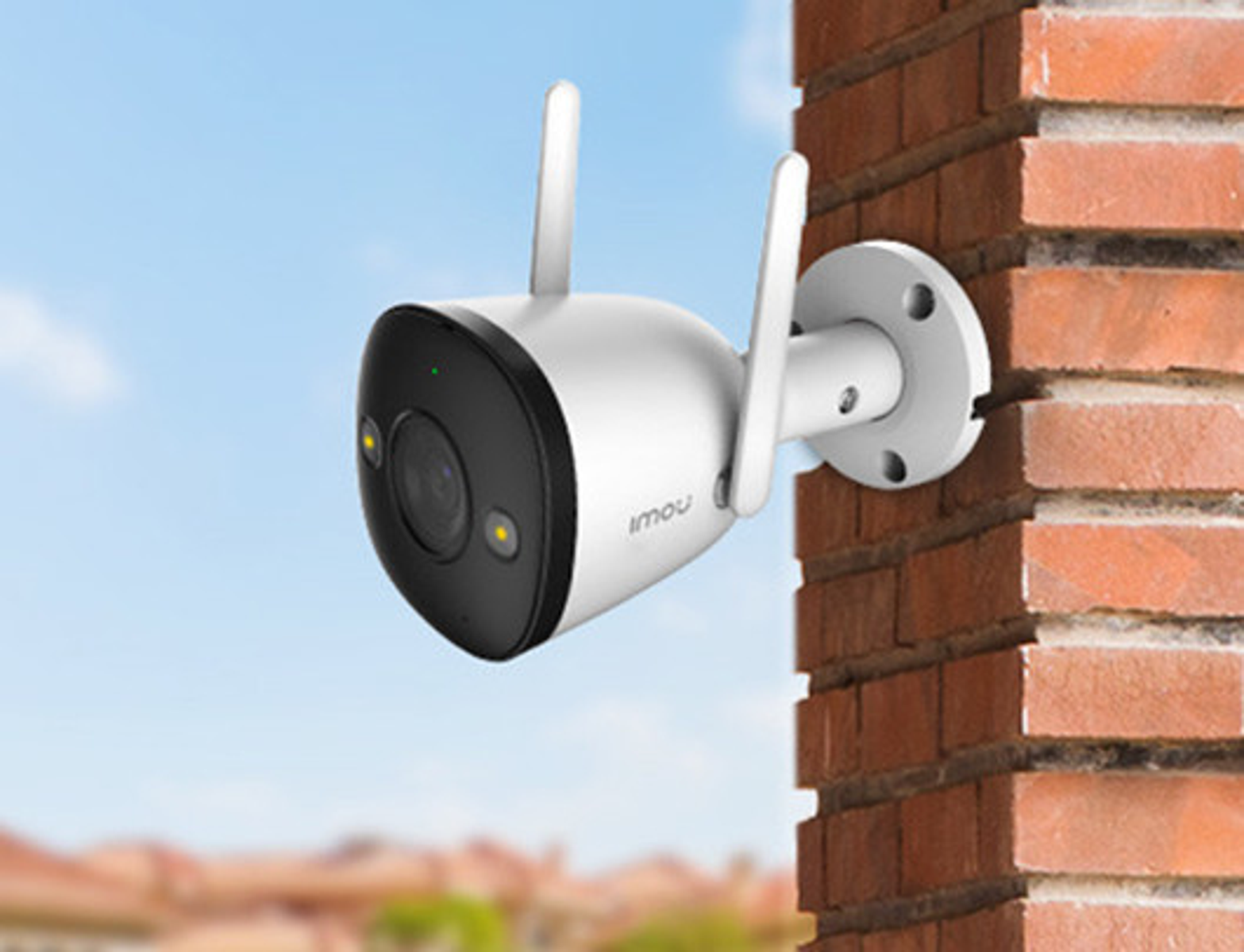 White security camera with two antennas, mounted on a brick wall, against a blue sky.