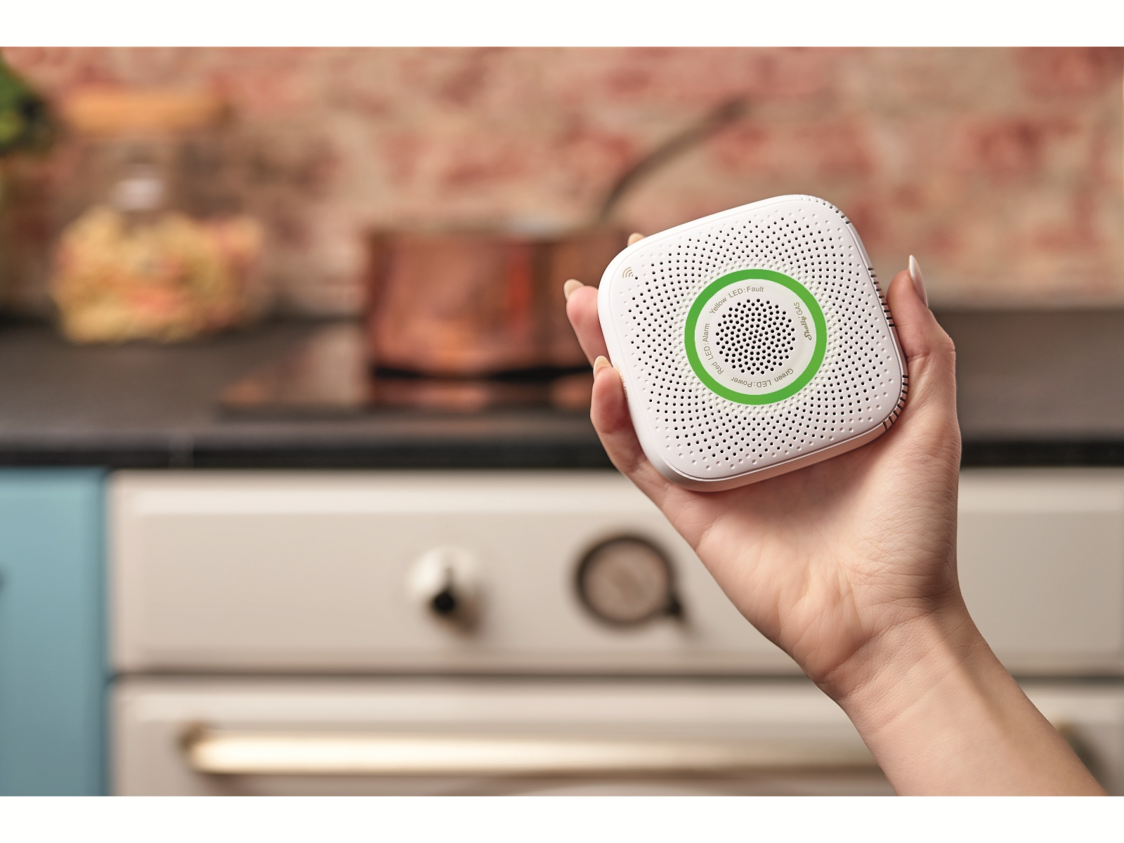 Dłoń trzymająca biały, kwadratowy czujnik gazu nad kuchenką w kuchni.