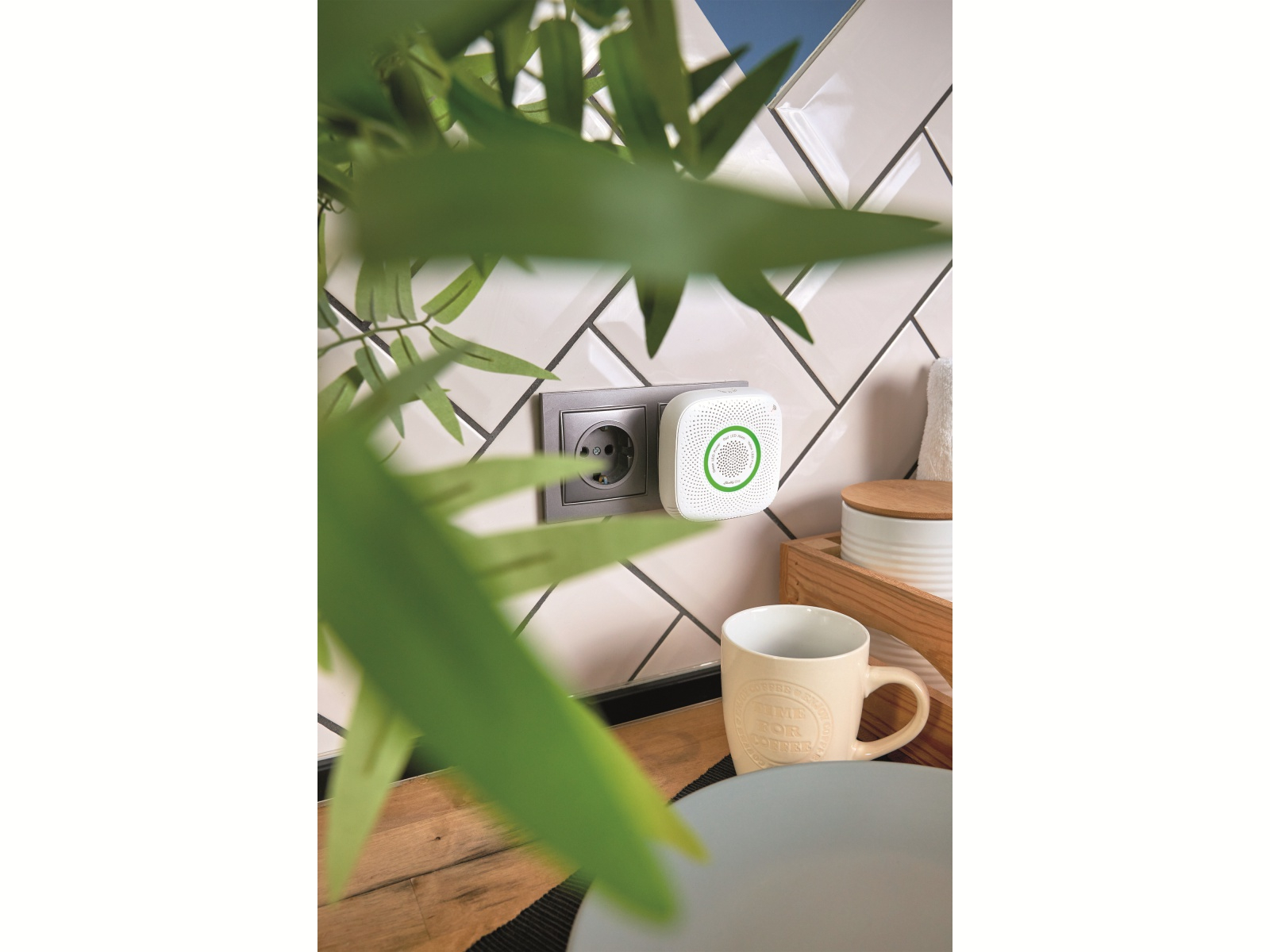 Biały czujnik gazu na ścianie obok gniazdka, filiżanka i talerz na stole.