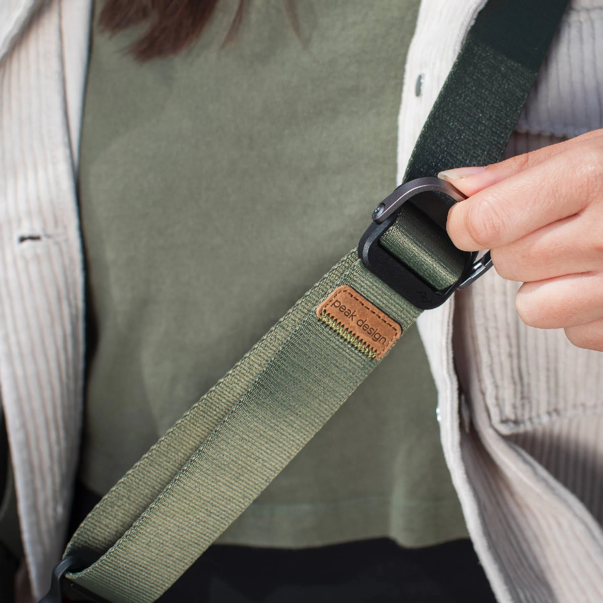 Zbliżenie zielonego paska do aparatu i dłoni regulującej klamrę. Beżowa koszula.