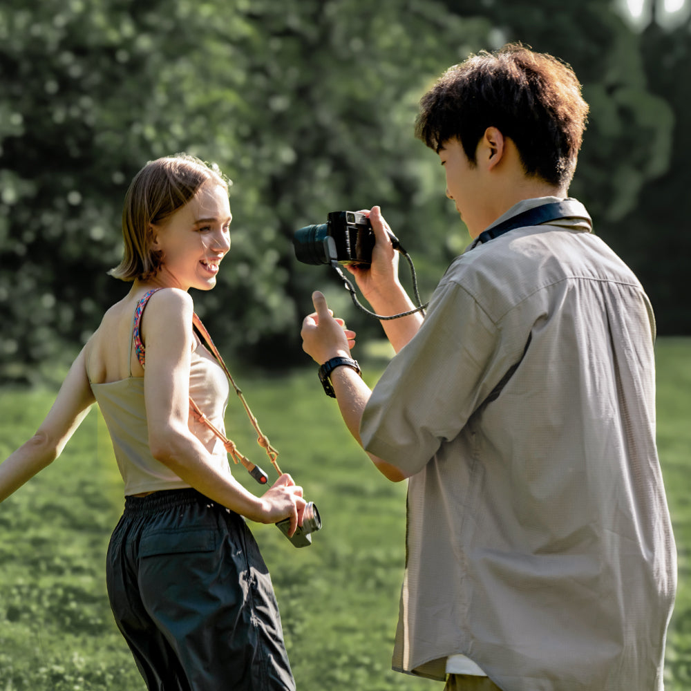 Un uomo scatta una foto a una donna sorridente in un parco. Erba verde e alberi sullo sfondo.