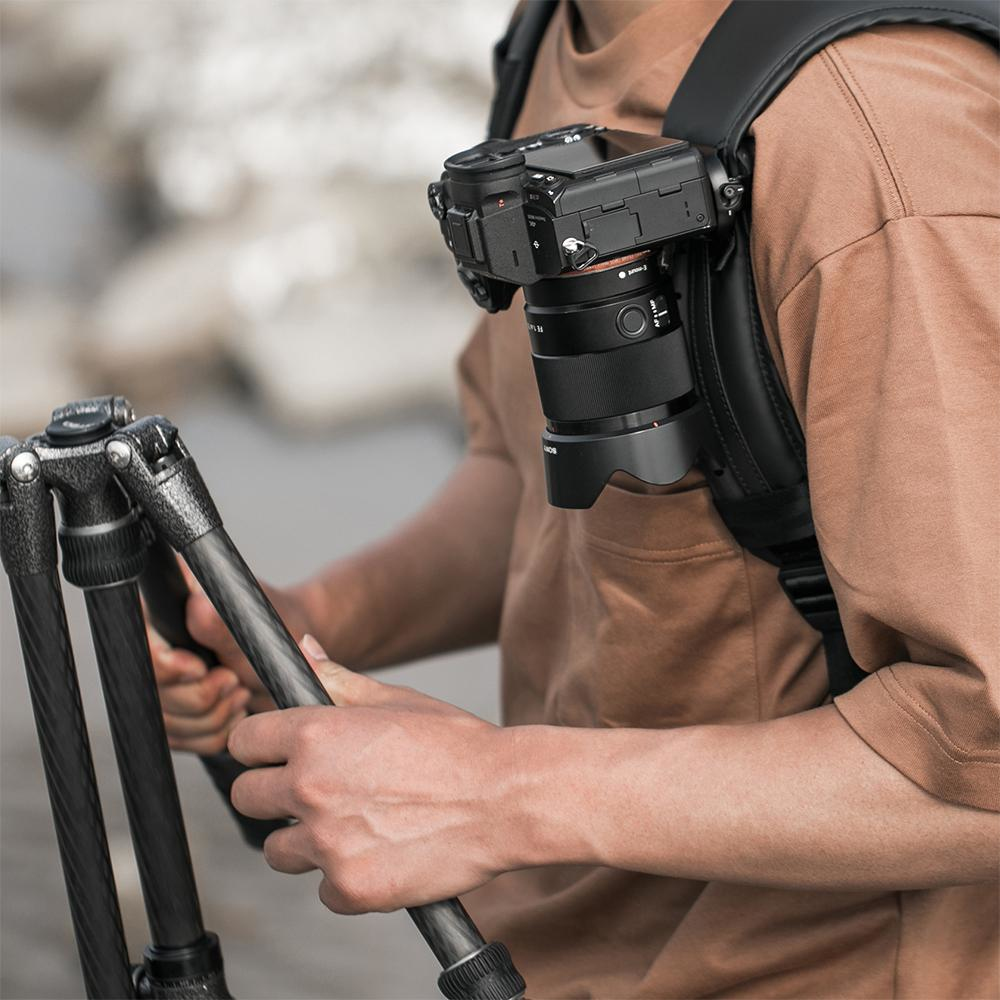 Mężczyzna trzymający statyw i aparat zamontowany na pasku, brązowa koszula, otoczenie zewnętrzne.