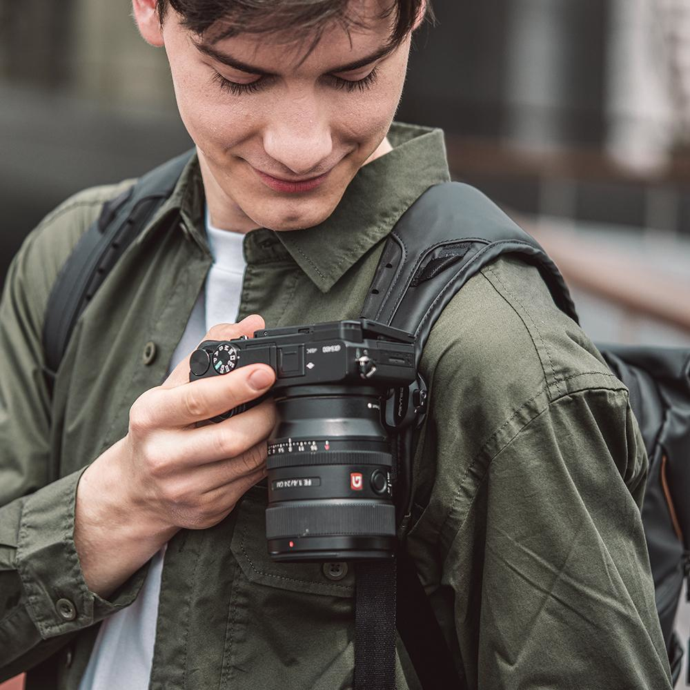 Młody mężczyzna patrzy na aparat zawieszony na pasku na ramieniu, uśmiechając się. Ma na sobie oliwkową koszulę.