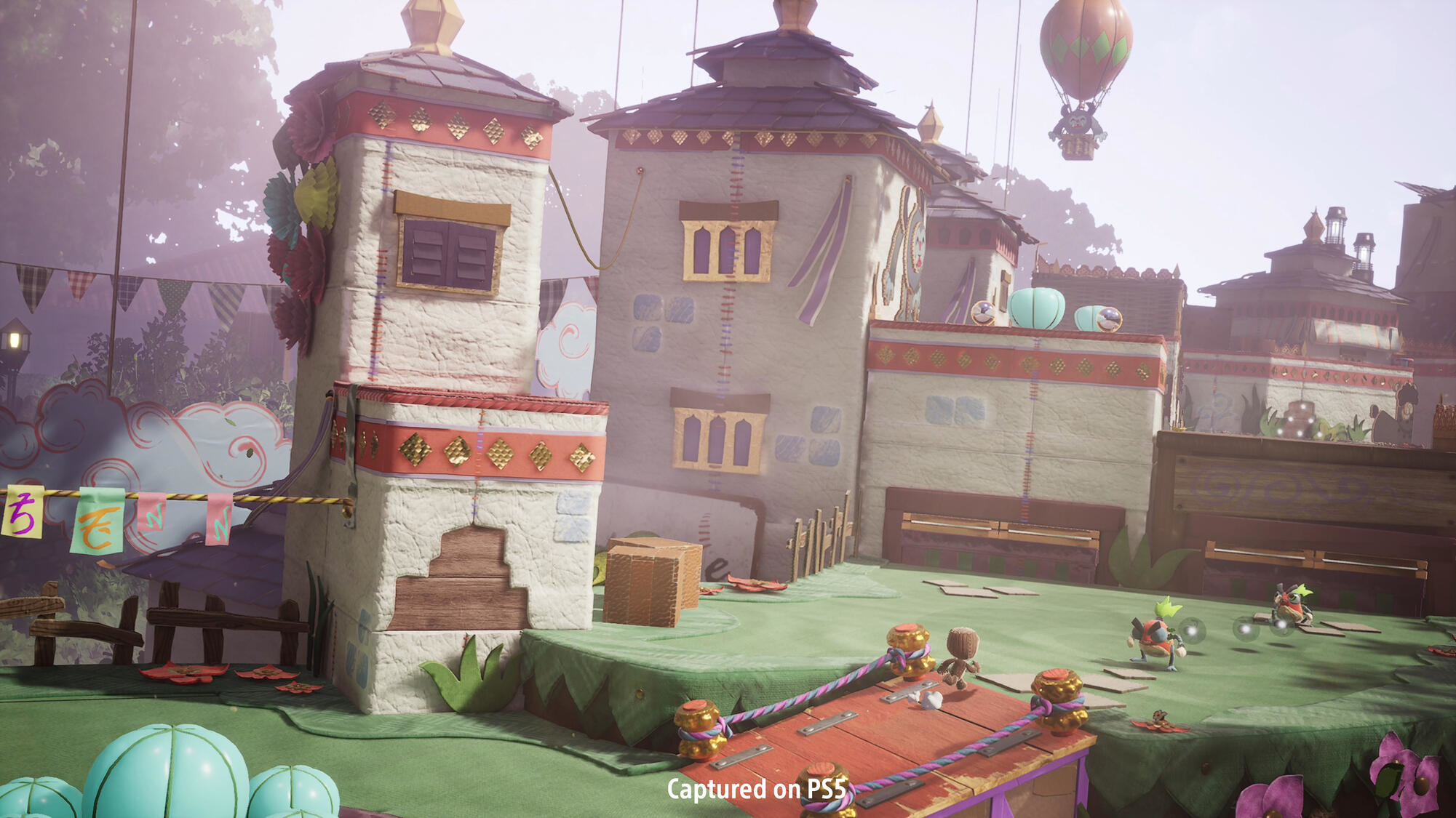 Scena z gry. Budynki, postacie i balon na ogrzane powietrze. Dzień, z lekkim, kolorowym stylem.