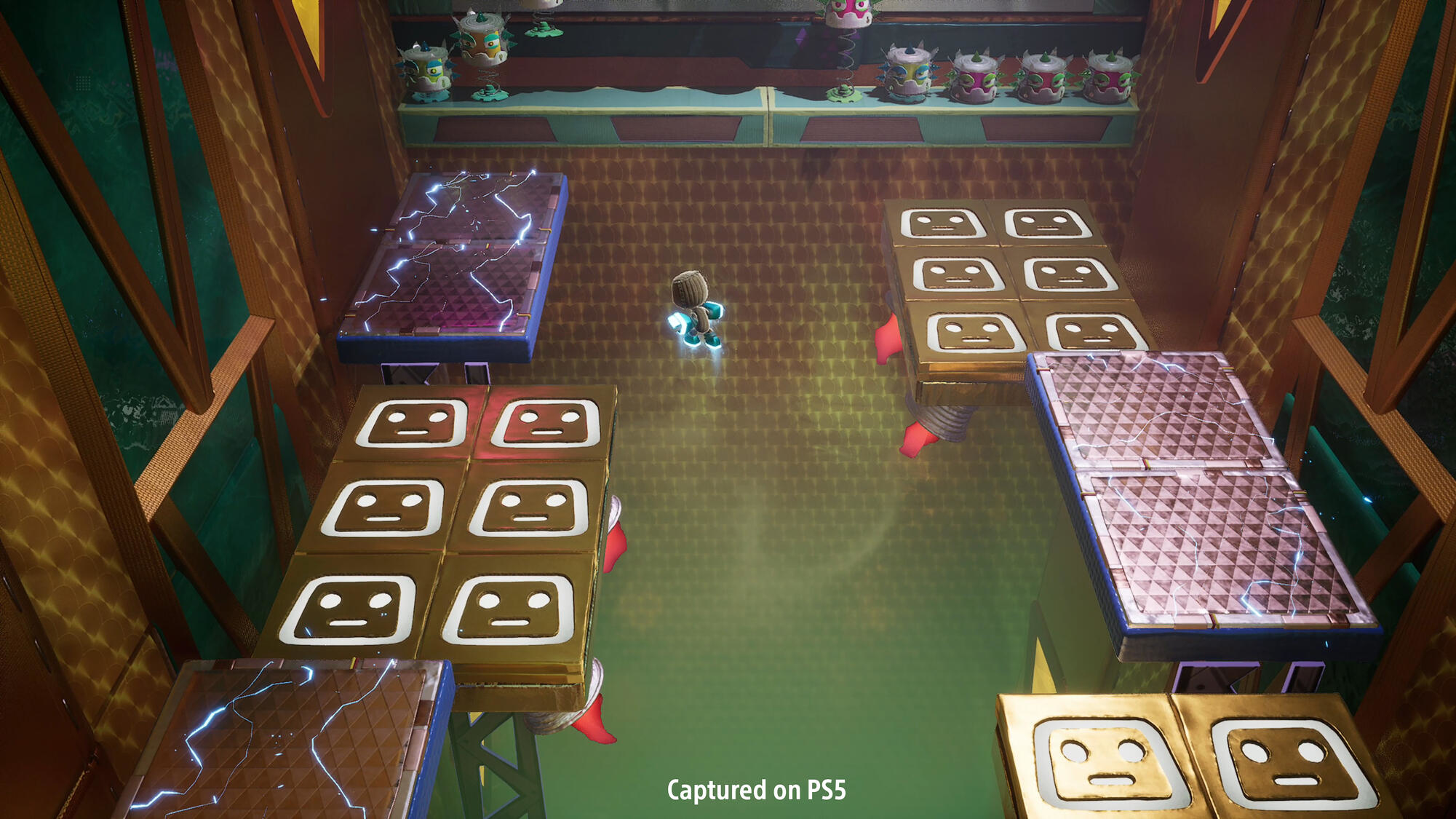 Postać w niebieskim kombinezonie stoi na platformie z płytkami w kształcie twarzy. Otoczenie jest wewnętrzne.