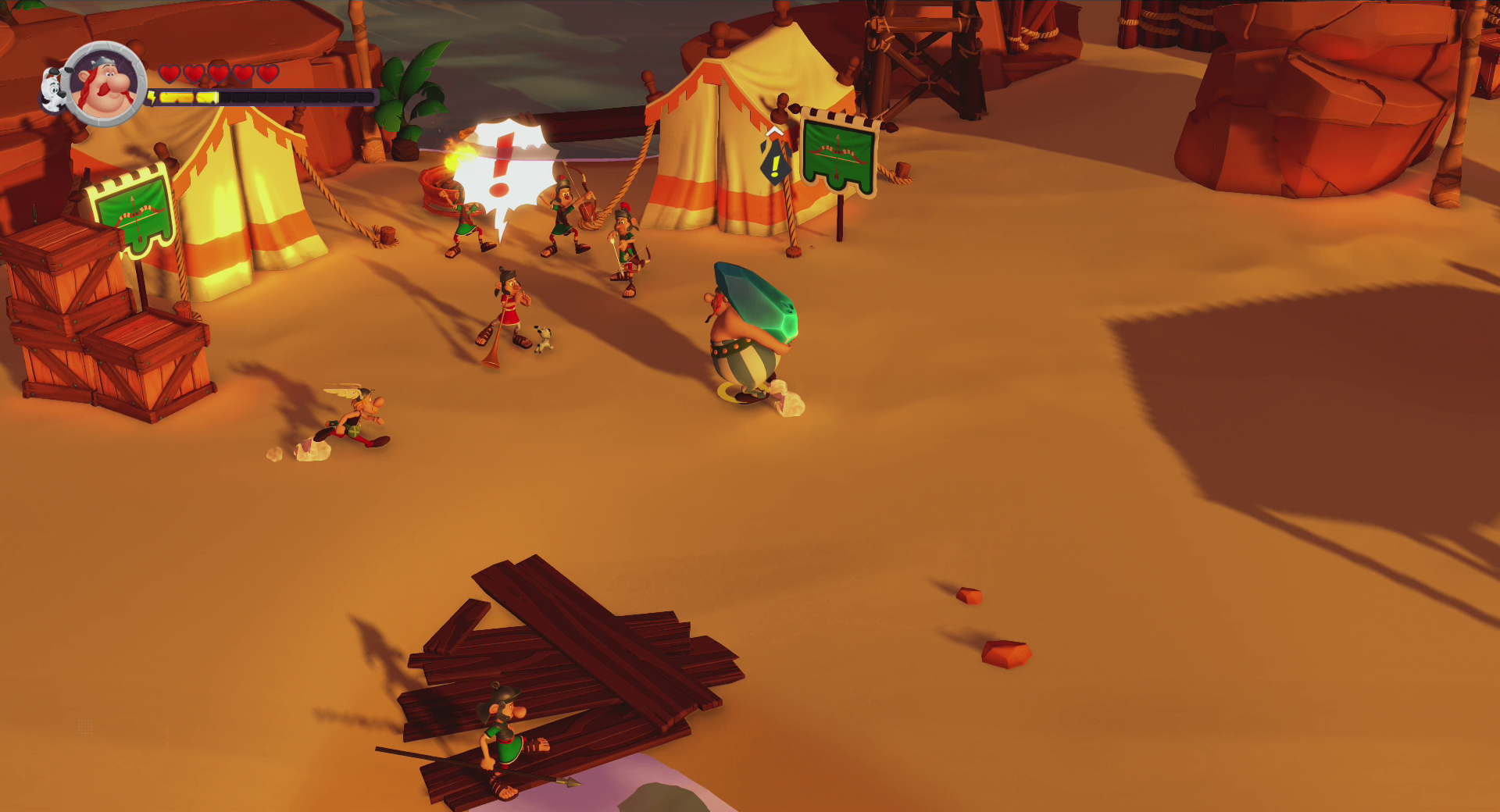 Scena del gioco Asterix. Diversi personaggi in un ambiente desertico. Casse di legno e tende sullo sfondo.