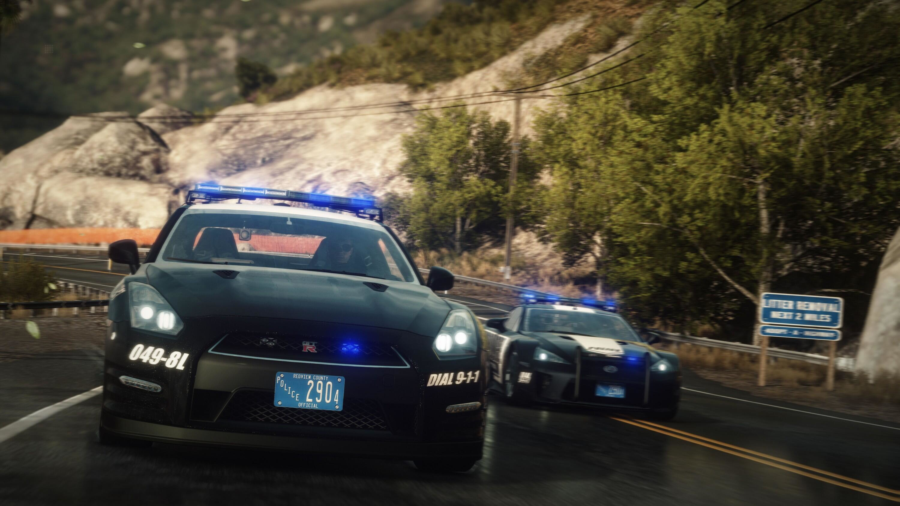 Due auto della polizia che corrono. Una è nera con luci blu. L'altra è bianca. Strada di montagna sullo sfondo.