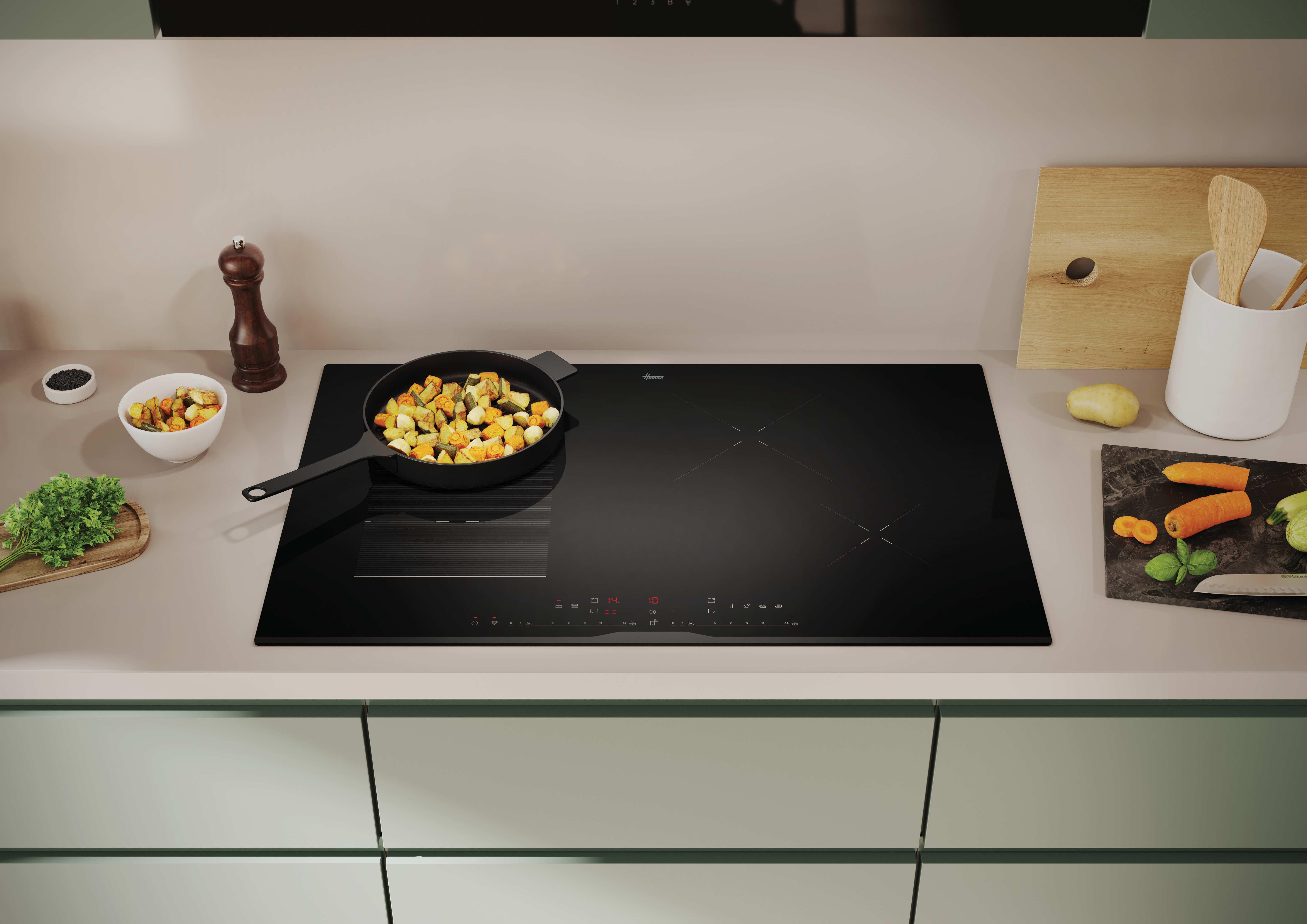 Piano cottura nero con cibo in padella. Sfondo mostra bancone, tagliere e utensili cucina.