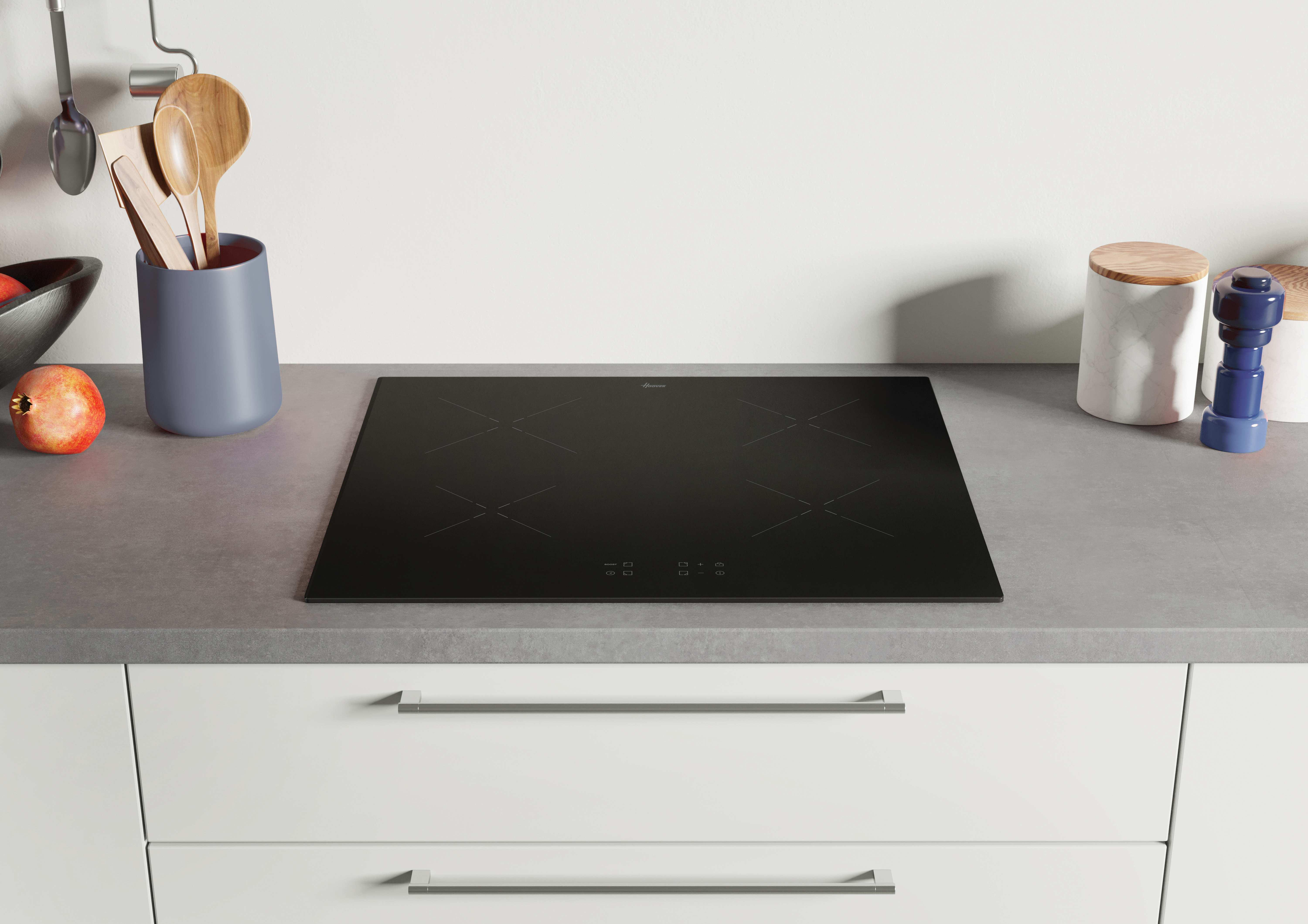 Piano cottura a induzione nero su piano grigio. Utensili da cucina e un melograno visibili.