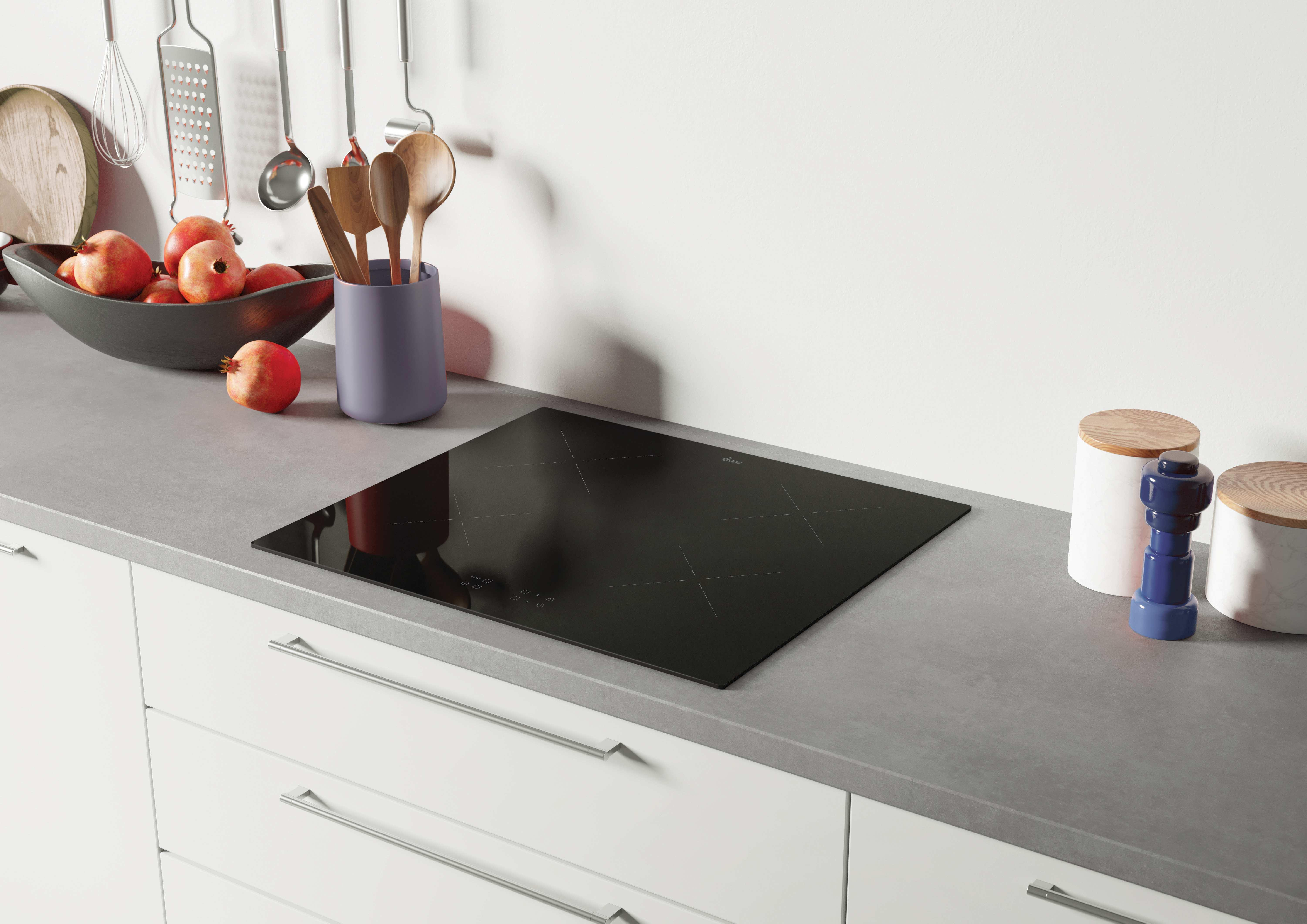 Piano cucina con piano cottura nero, utensili, ciotole di frutta e barattoli. Sotto, mobili bianchi.