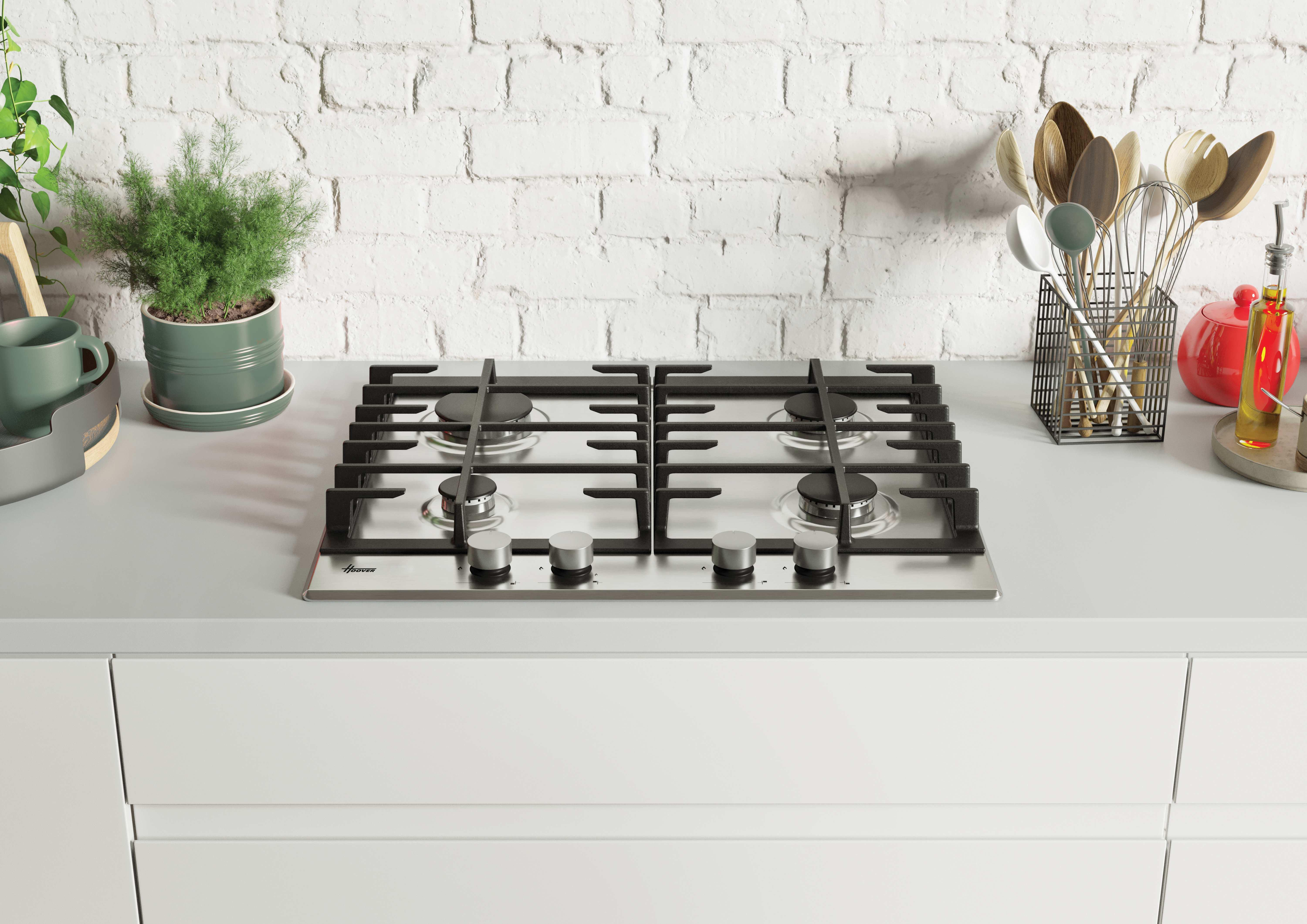 Encimera de cocina con placa de gas, plantas, utensilios y pared de ladrillo al fondo. Armarios blancos debajo.