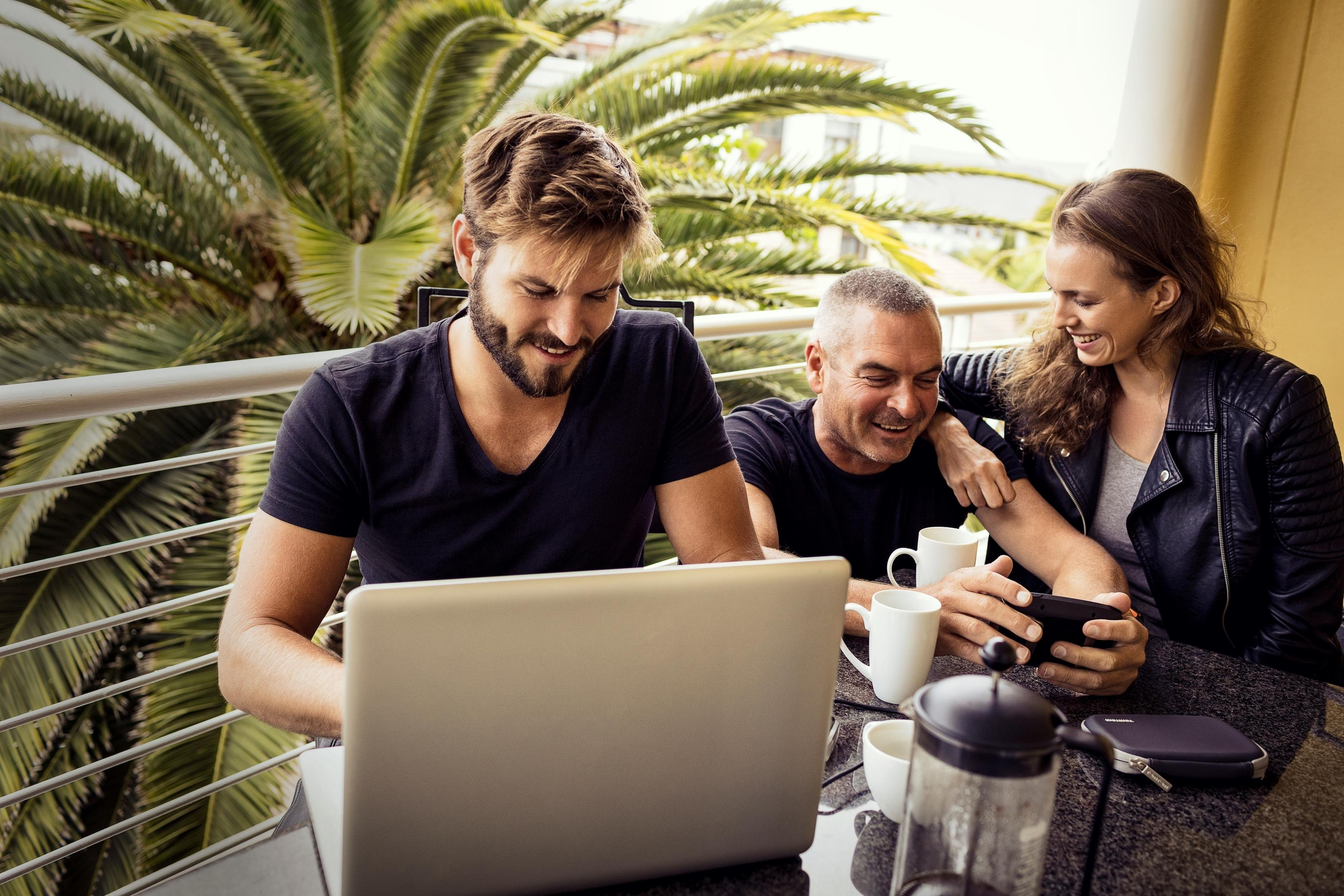 Drei Personen auf einem Balkon, benutzen Laptop und Smartphone, trinken aus Tassen, Palmen im Hintergrund.