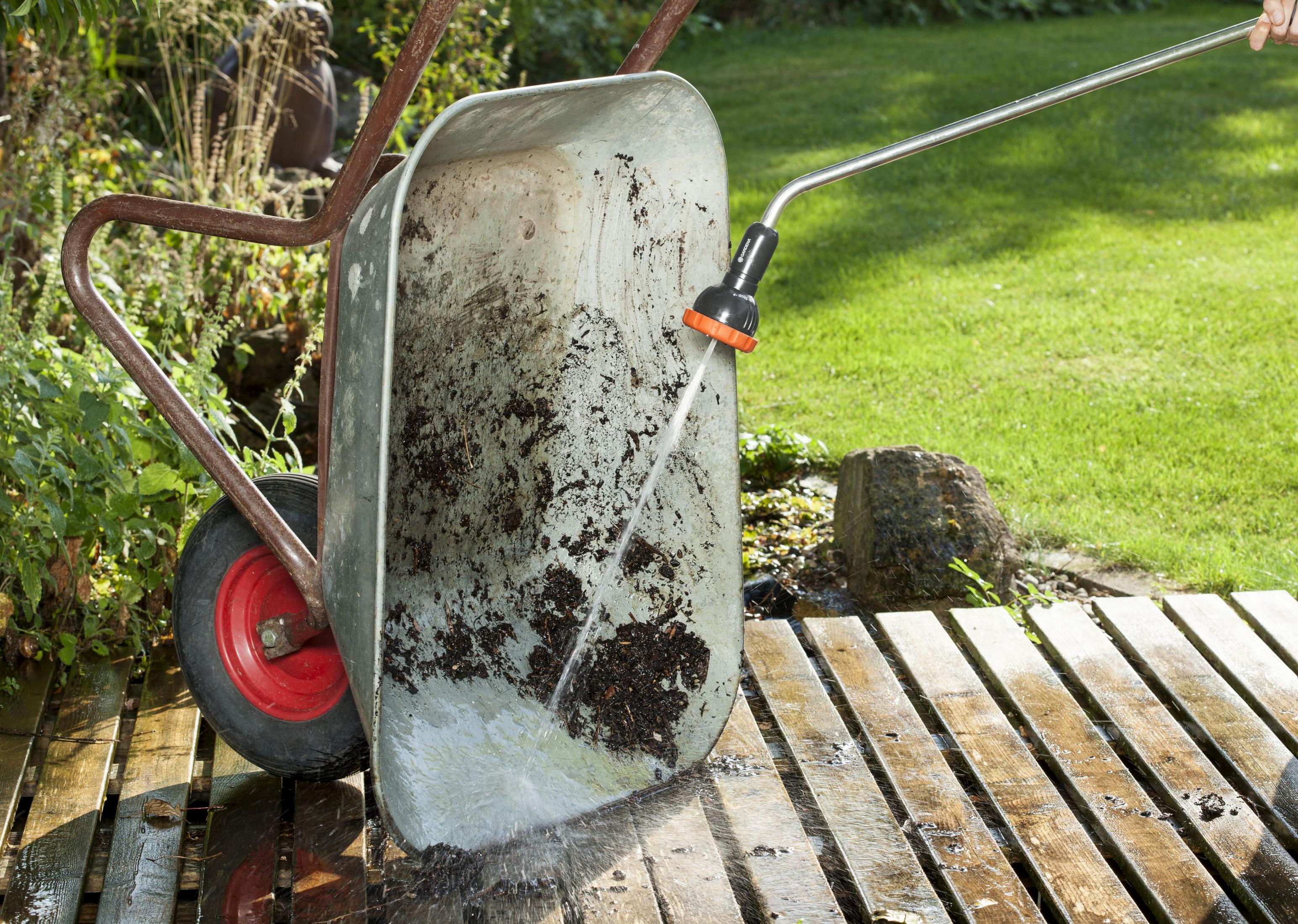 Eine Person sprüht Wasser aus einem Schlauch in eine schmutzige Schubkarre auf einer Holzterrasse im Garten.