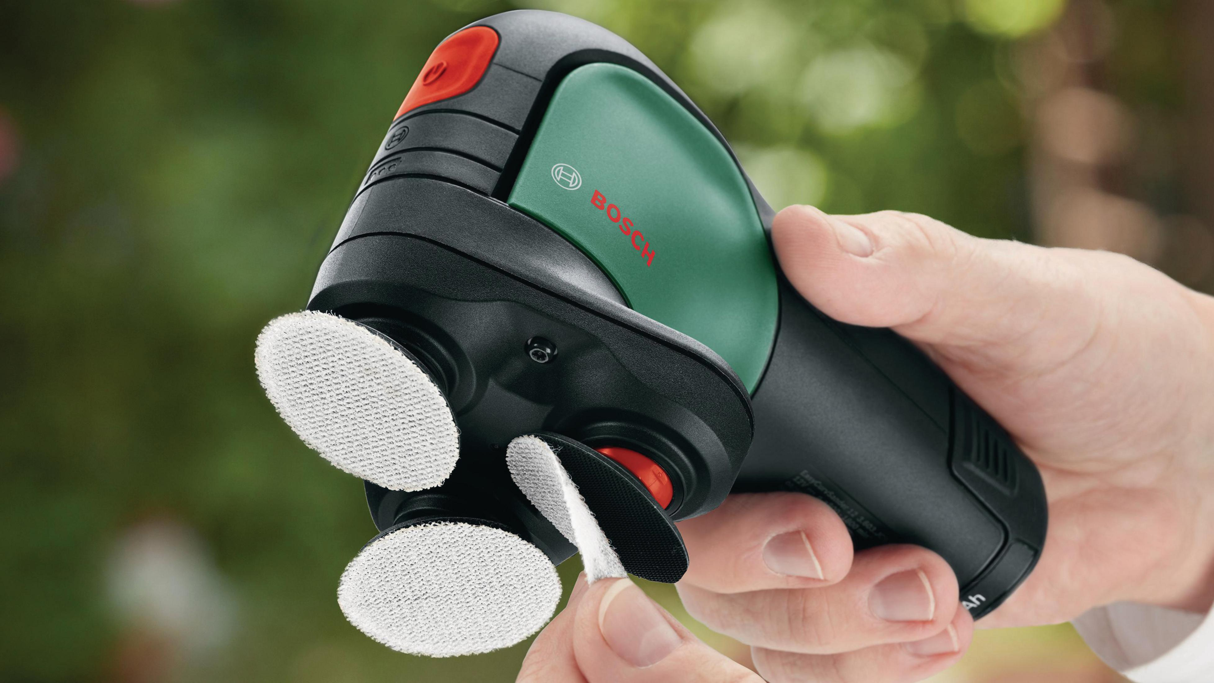 Une main utilisant une ponceuse multiple Bosch avec des éléments de couleur verte, noire et rouge.
