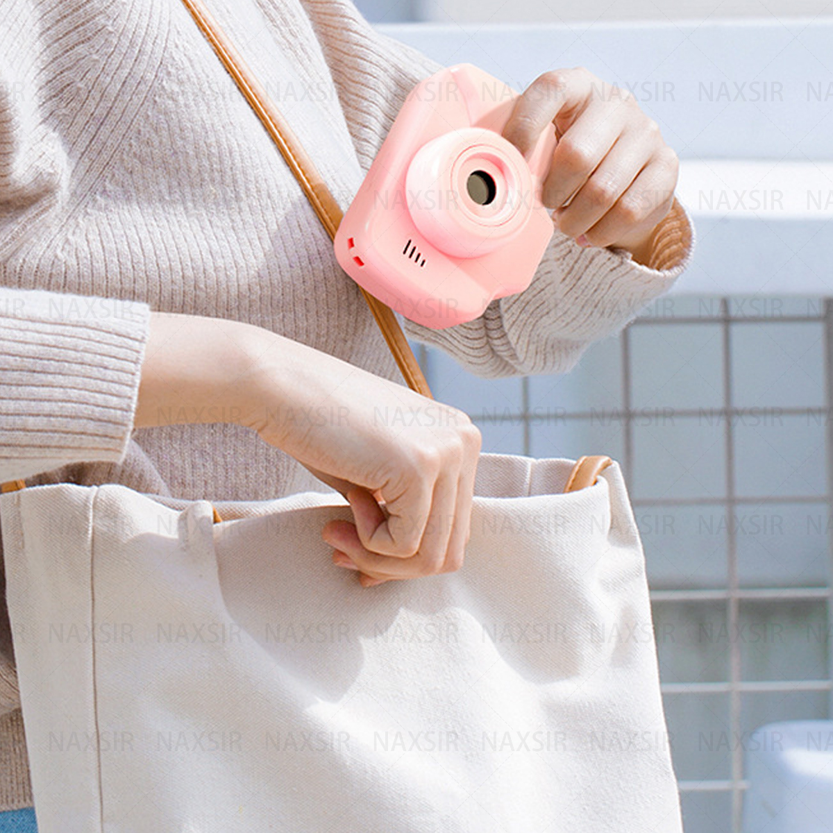 Una fotocamera rosa tenuta da qualcuno davanti a una borsa bianca. La fotocamera ha un obiettivo e dei pulsanti.