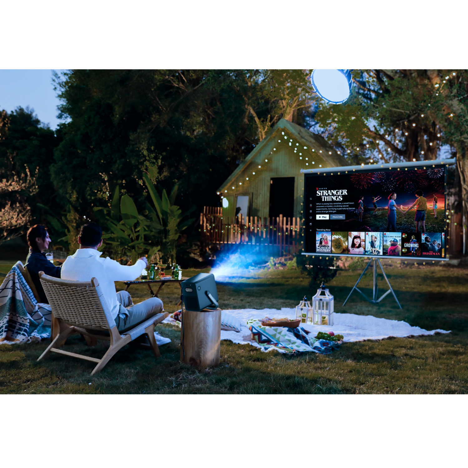 Escena de cine al aire libre con proyector, pantalla y personas viendo una película.