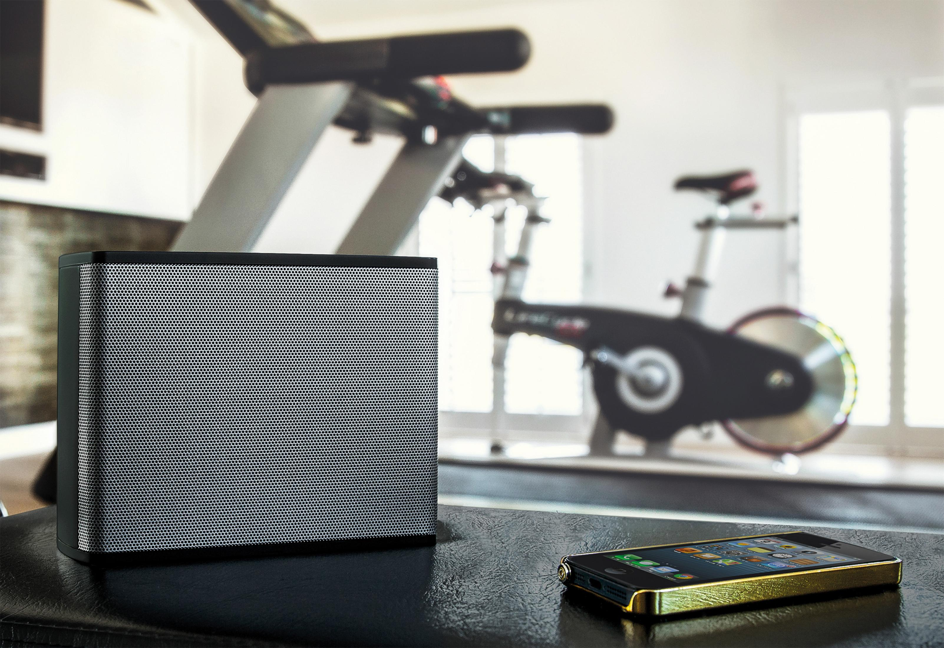 Zwarte luidspreker en een gouden iPhone op een tafel in een sportschool. Een loopband en een hometrainer staan op de achtergrond.