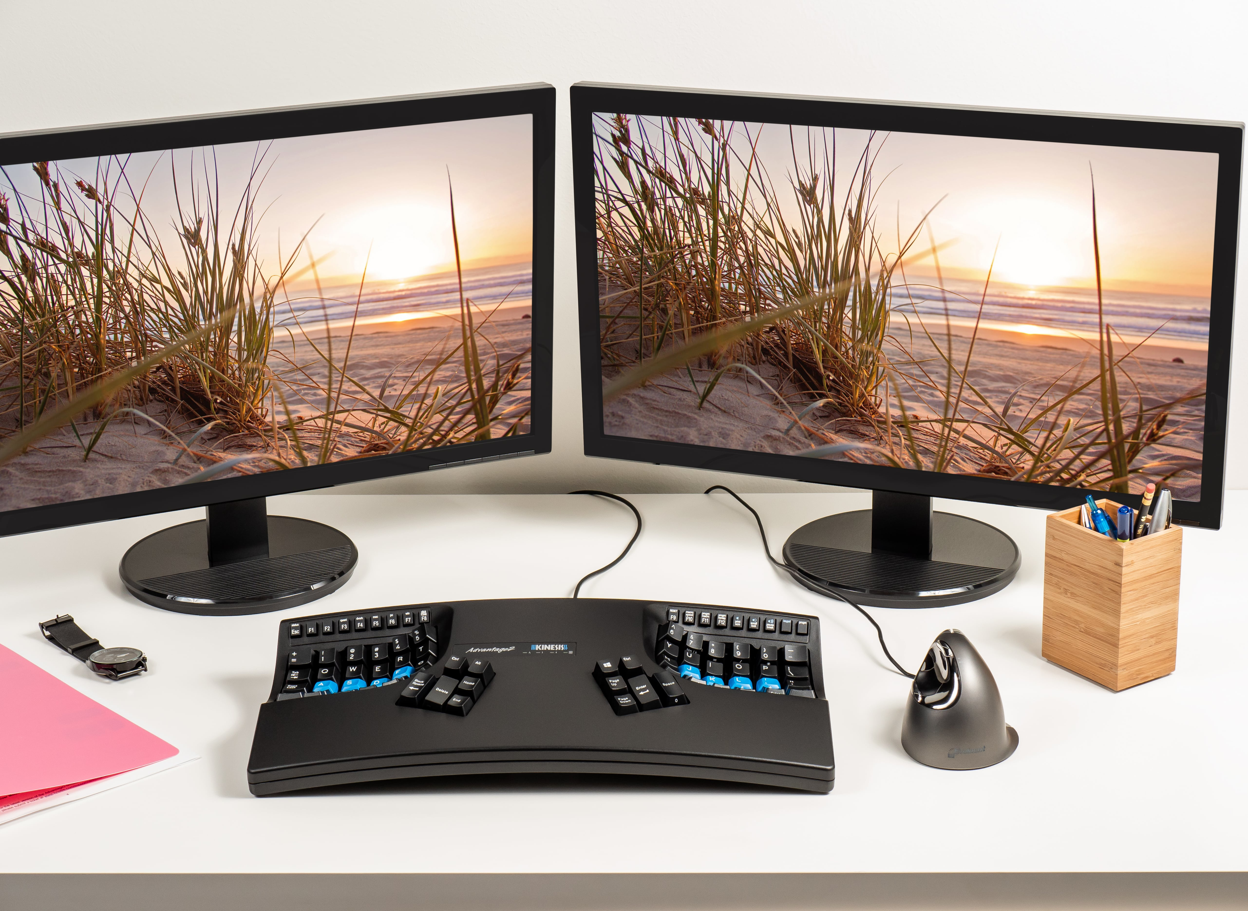Tastiera ergonomica con due monitor che mostrano una scena di spiaggia. Portapenne in bambù e orologio presenti.