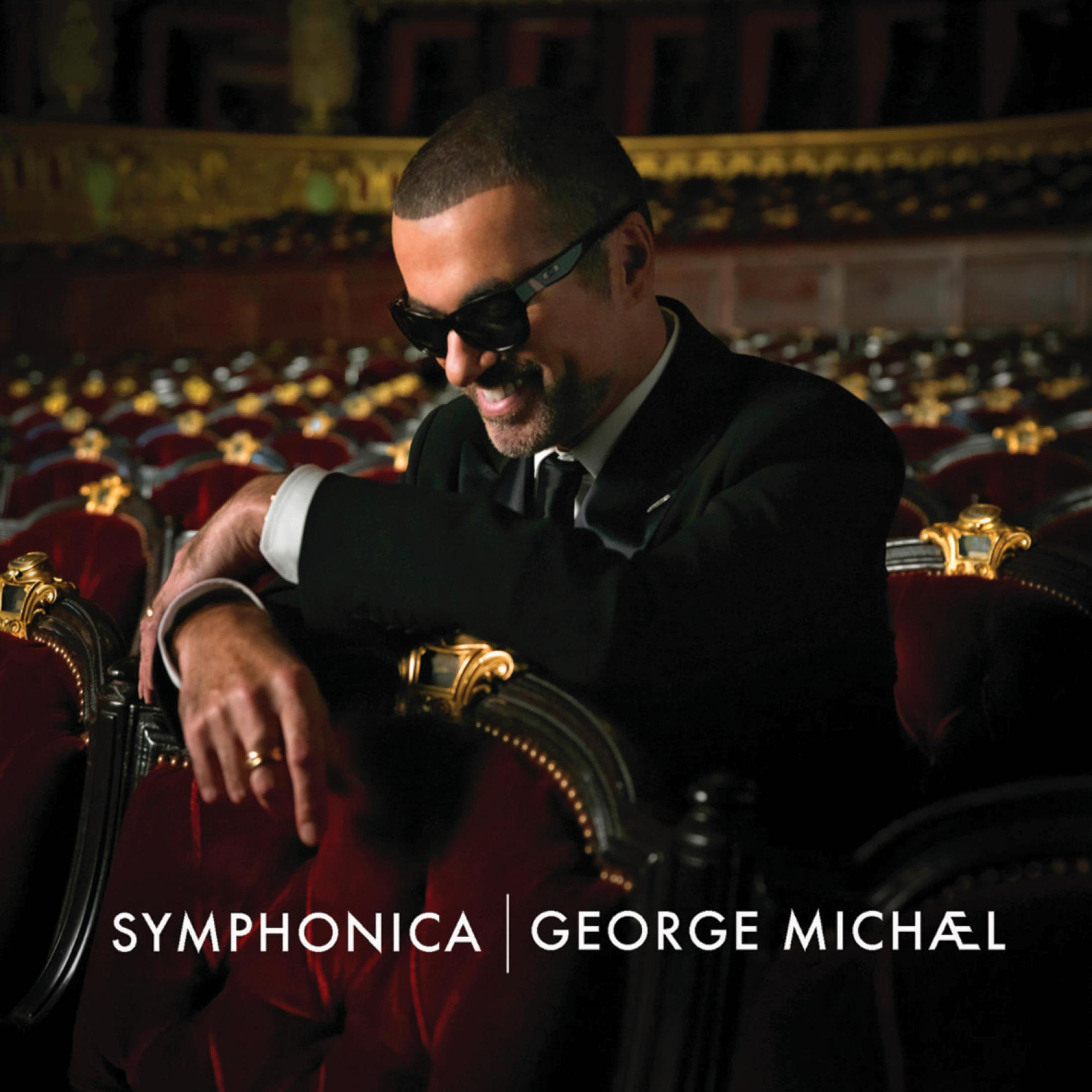 George Michael in abito e occhiali da sole, seduto su una poltrona di velluto rosso, sorridente.