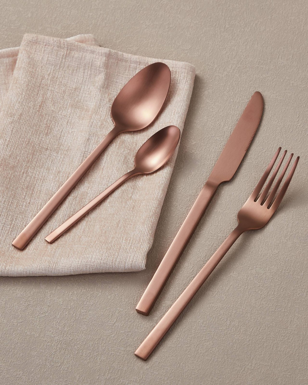 Quattro utensili color oro rosa sono su un tovagliolo beige. Un cucchiaio, un cucchiaino, un coltello e una forchetta, su sfondo beige.