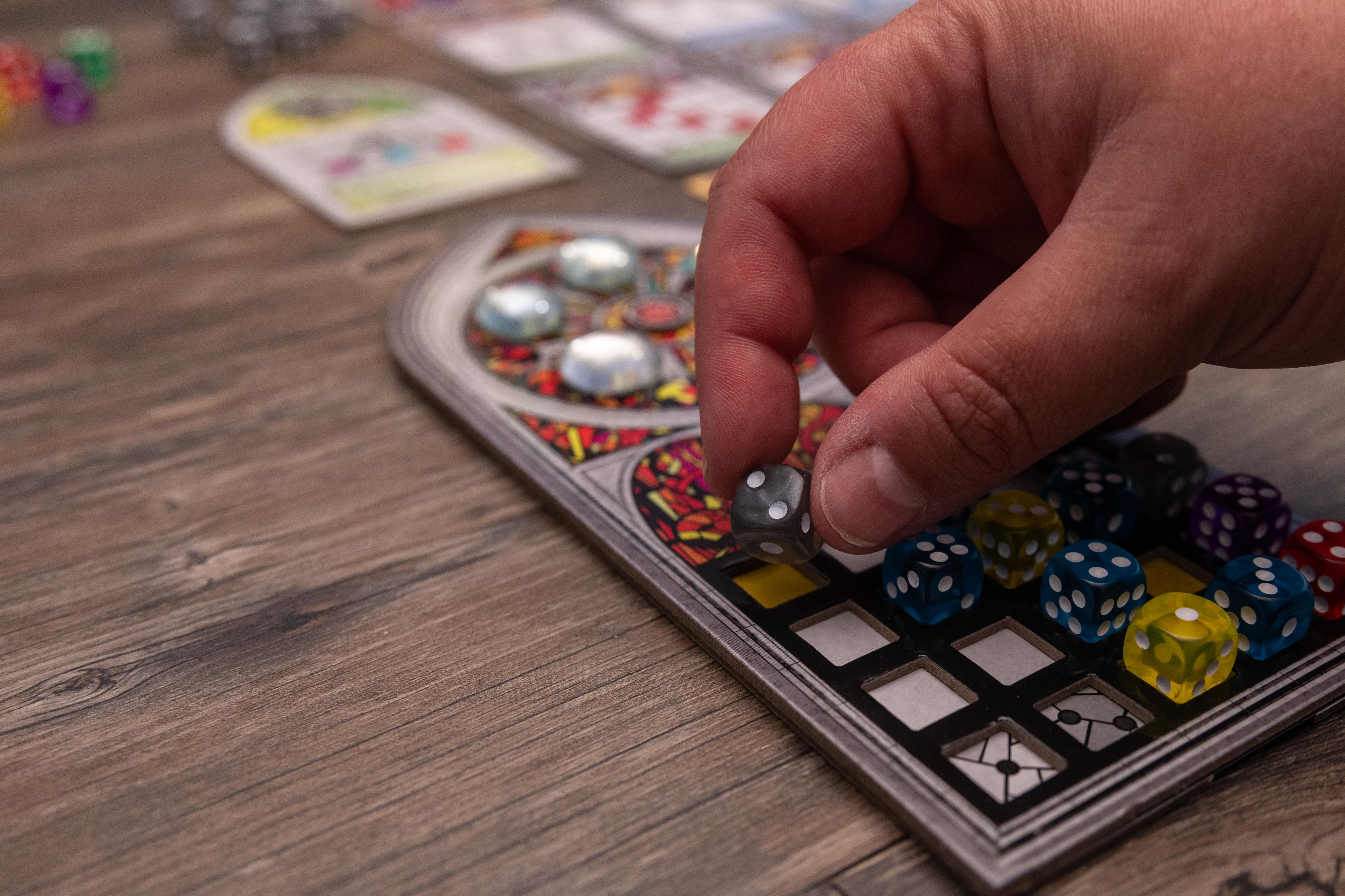Eine Hand, die einen Würfel auf ein Brettspiel legt. Das Brett hat ein Buntglasfenster-Design.