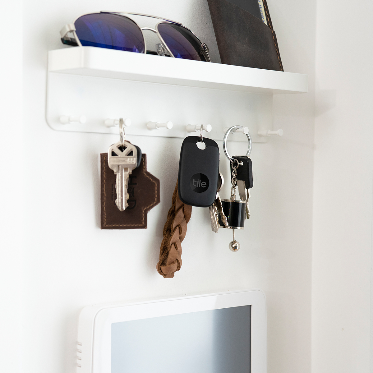 Klucze, okulary przeciwsłoneczne i lokalizator Tile wiszące na półce na ścianie. Biała ściana, biała półka.