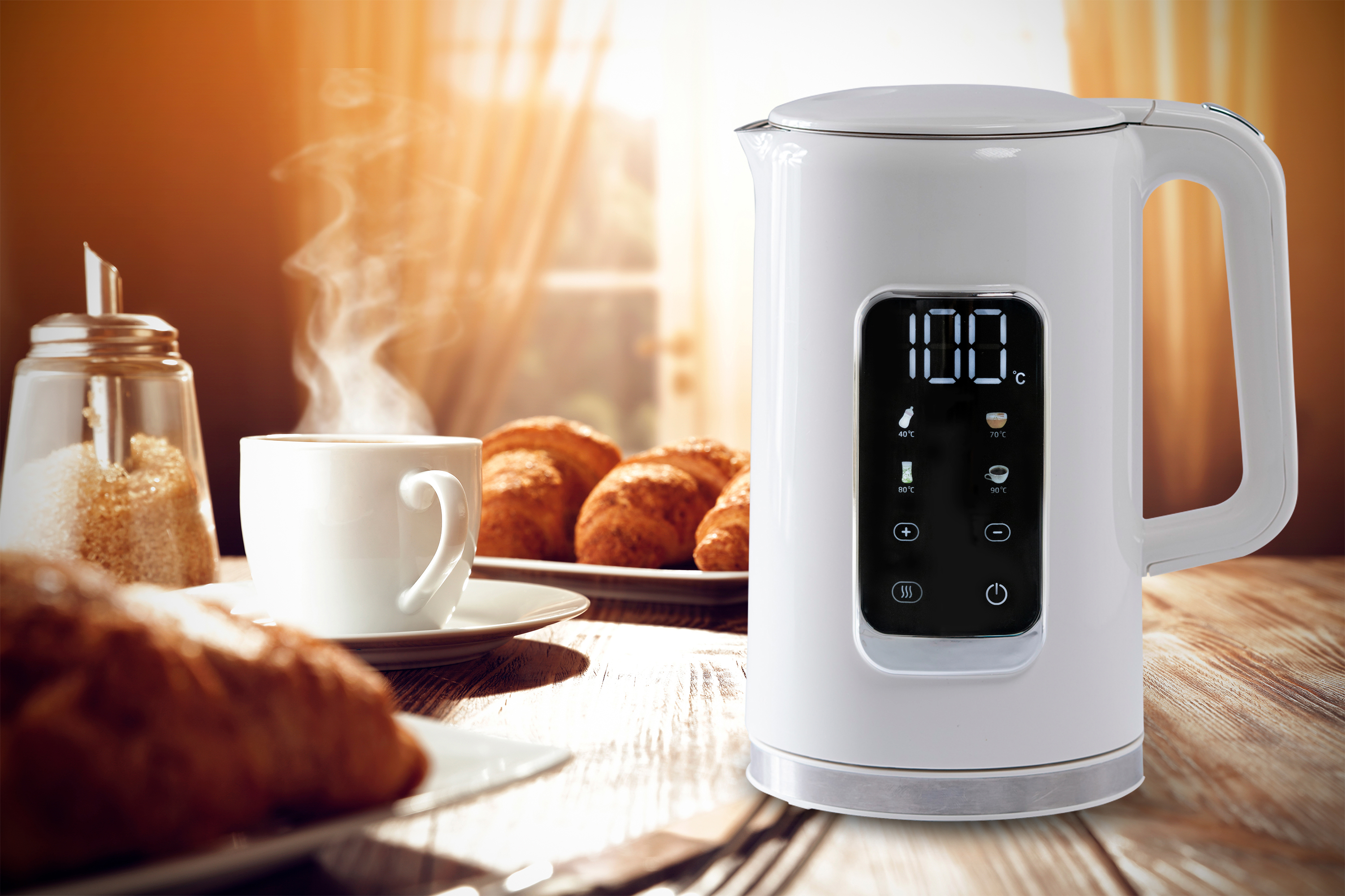 Biały czajnik elektryczny z produktami śniadaniowymi na stole. Pokazuje 100°C.
