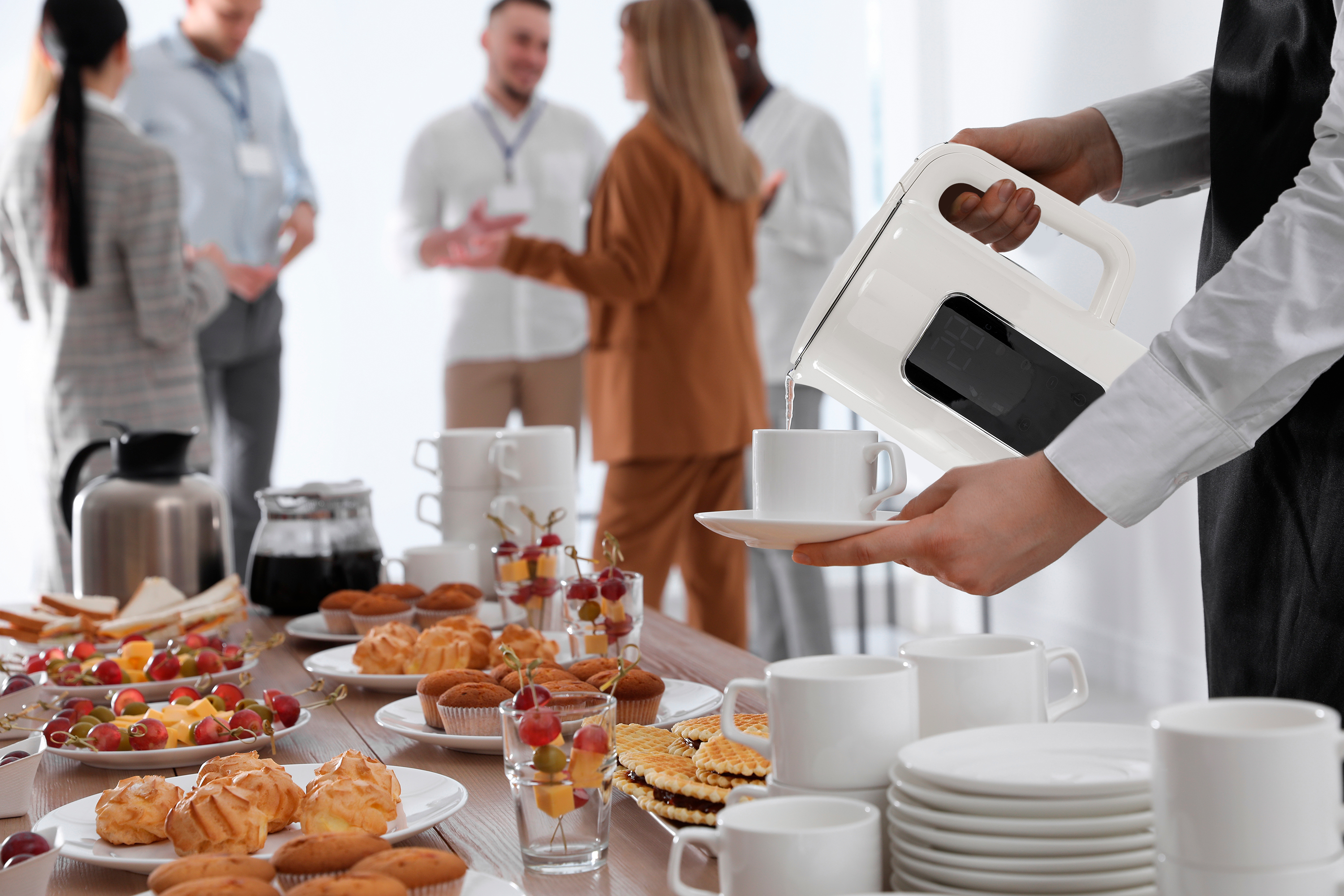 Osoba nalewa wodę z białego czajnika do filiżanki. Ludzie stoją w tle.
