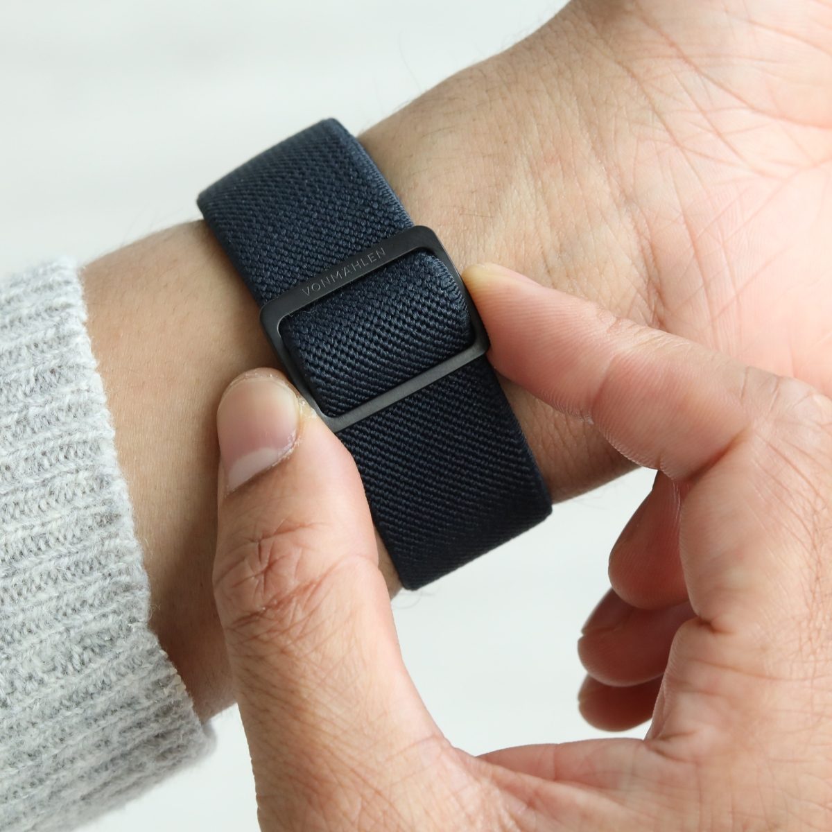 Una mano che indossa un cinturino blu scuro con fibbia nera. Il polso è coperto da un maglione grigio.