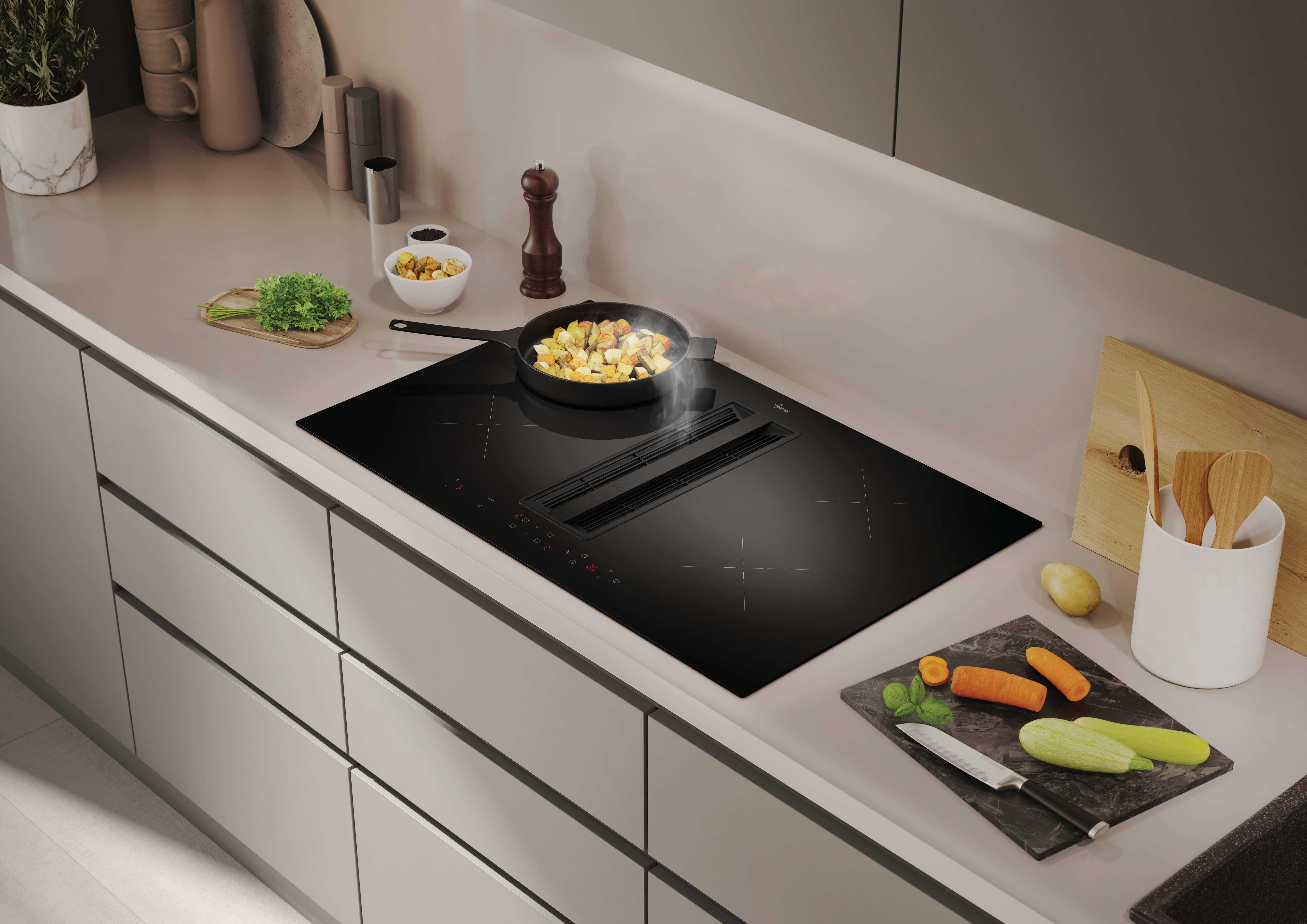 Escena de cocina con vitrocerámica negra, sartén con comida y verduras frescas en una tabla de cortar.