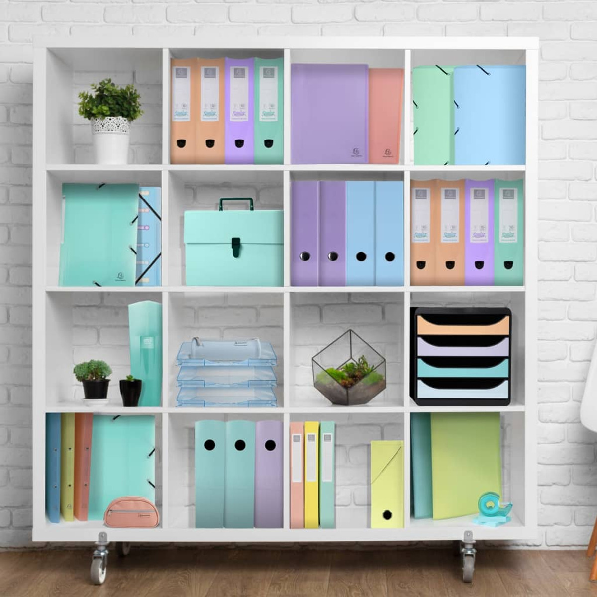 Biała jednostka półkowa z segregatorami, folderami i organizerami w różnych kolorach, na tle ceglanej ściany.