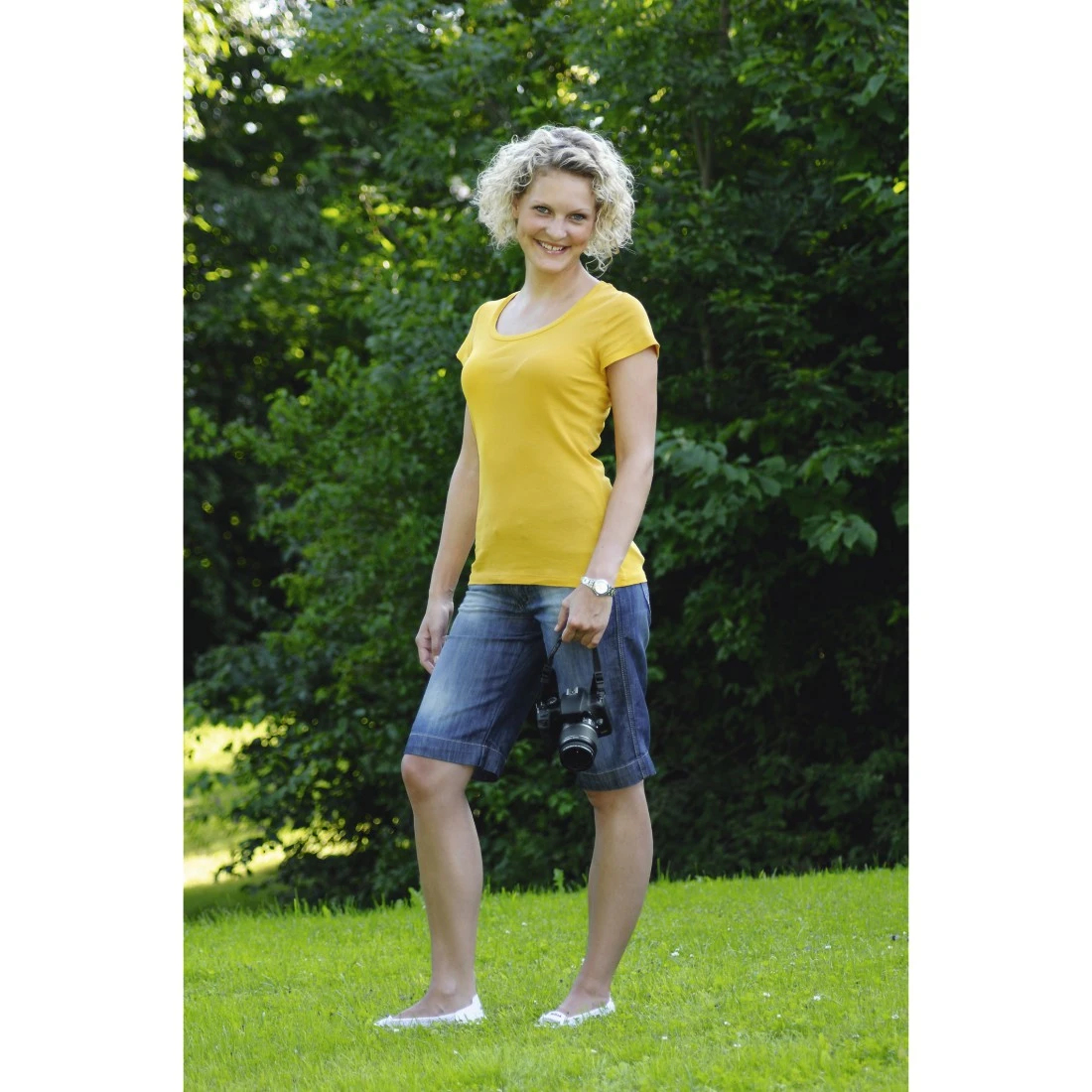 Donna bionda con maglietta gialla e pantaloncini di jeans che tiene una fotocamera.