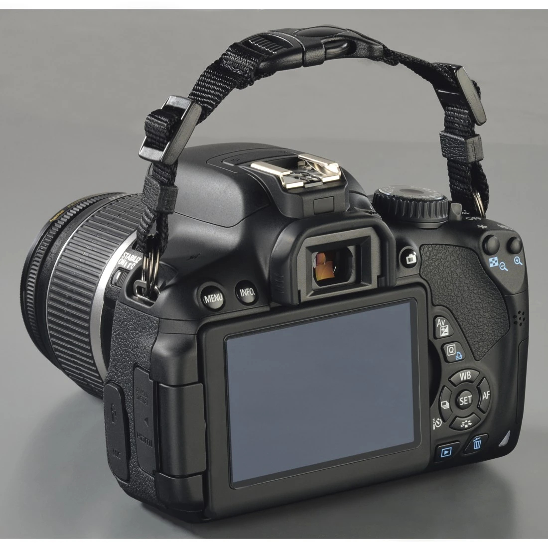 Fotocamera DSLR nera con cinturino, vista posteriore, su superficie grigia.