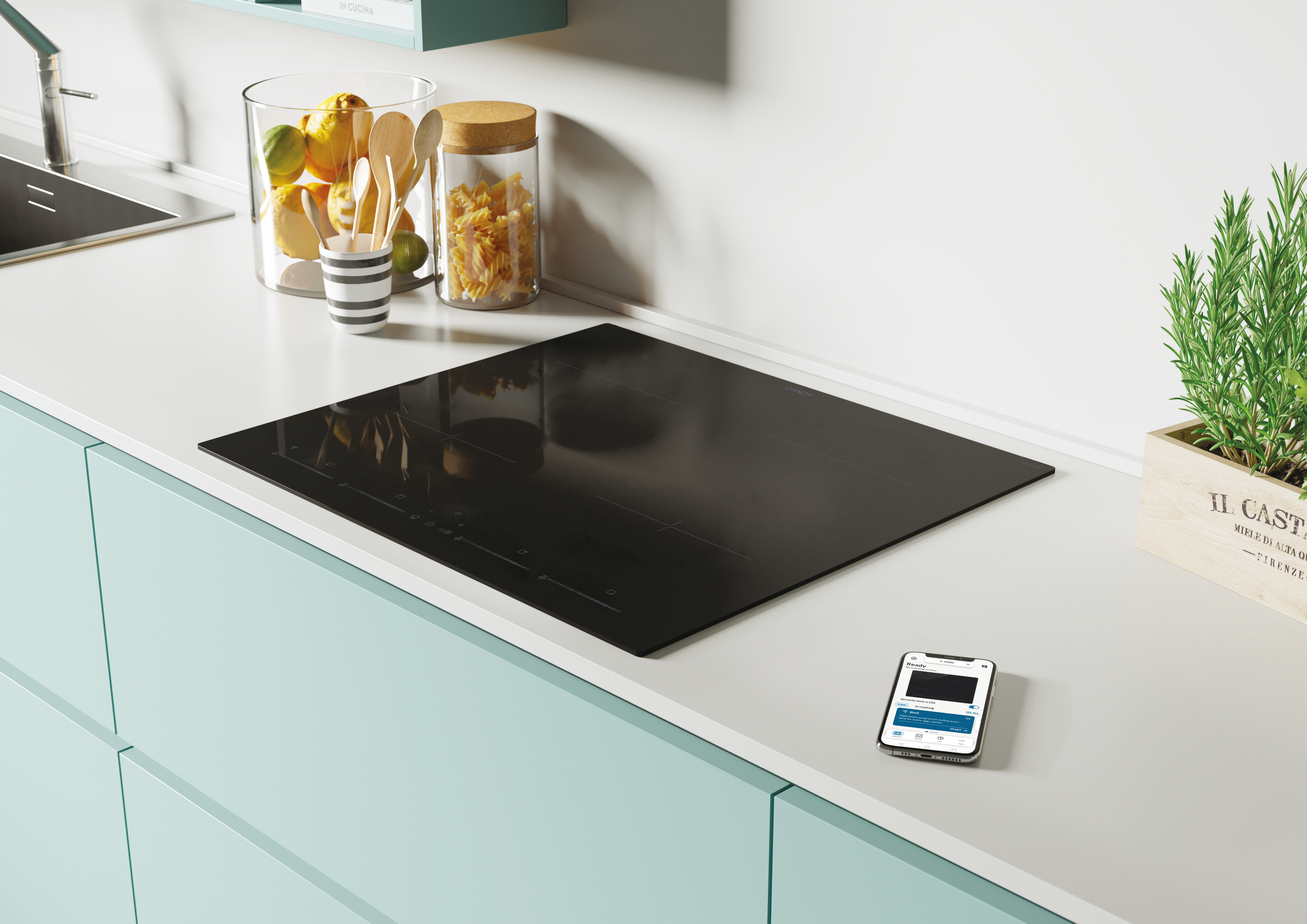 Czarna płyta indukcyjna na blacie. Słoik z makaronem, szklanka z drewnianymi łyżkami i smartfon.