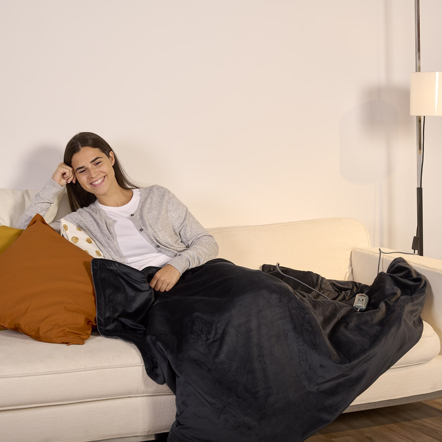 Kobieta na kanapie pod kocem. Lampa i poduszki są w tle.