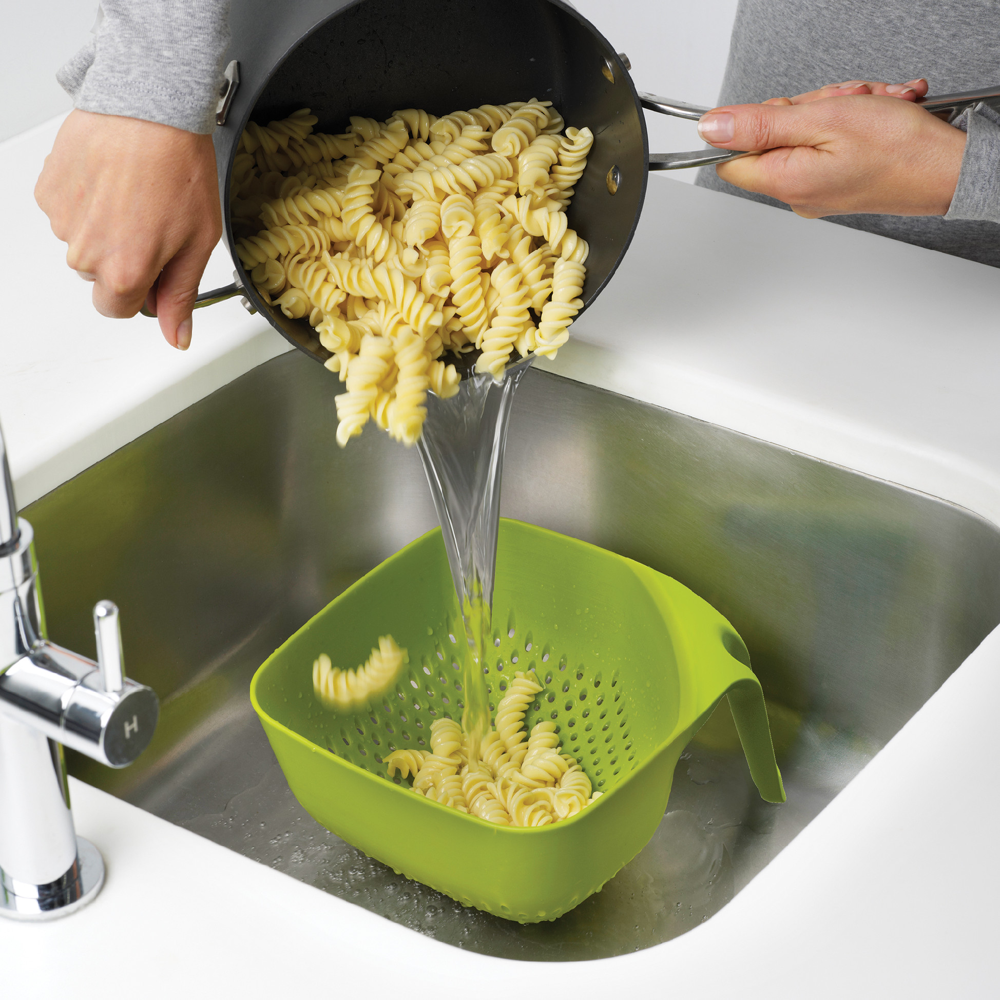 Pasta vertiéndose de olla negra a escurridor verde sobre un fregadero acero inoxidable.