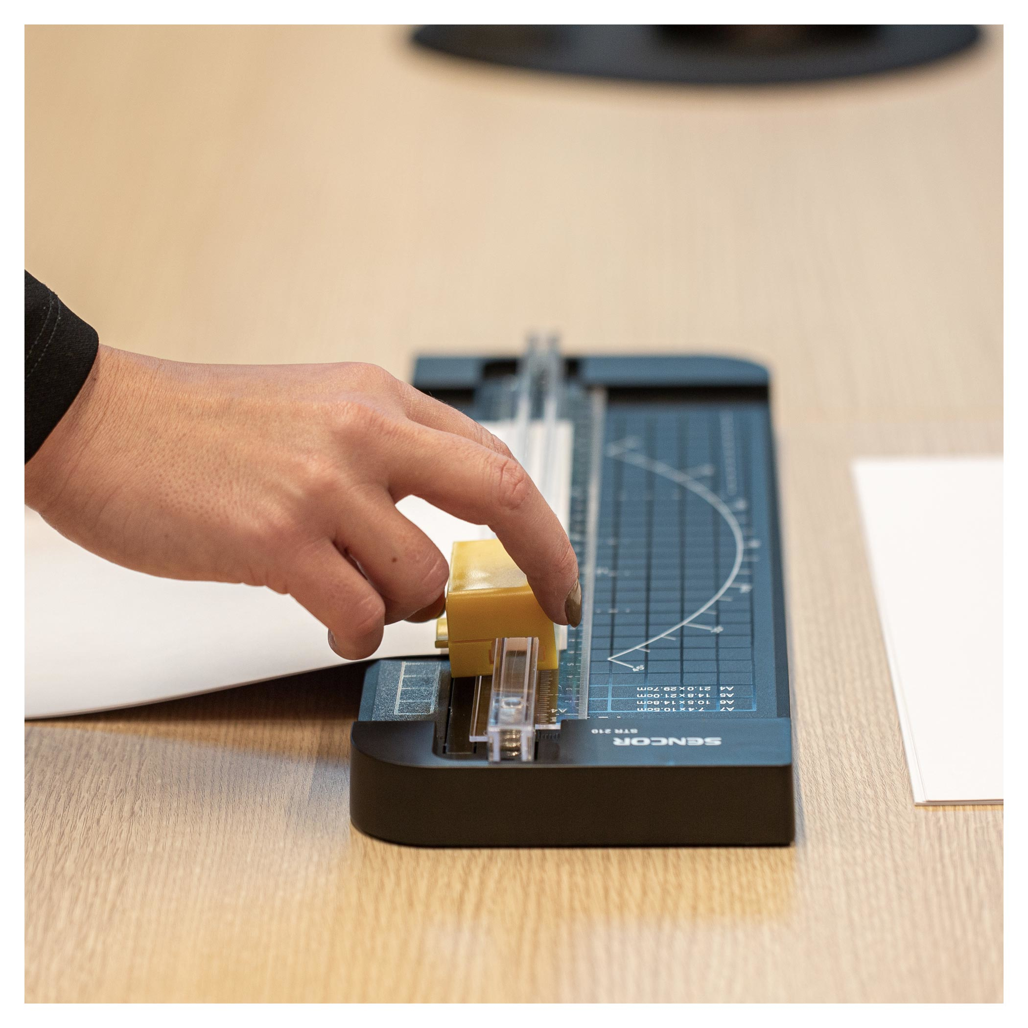 Ręka używa gilotyny do papieru na białej kartce, przed nią żółty element.