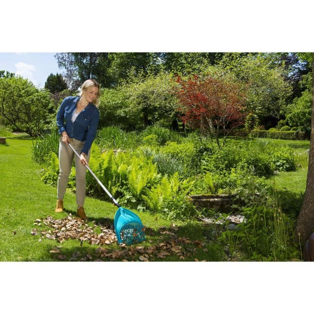Eine Frau harkt Laub in einem Garten. Sie benutzt einen türkisfarbenen Laubrechen. Grünes Gras und Bäume im Hintergrund.