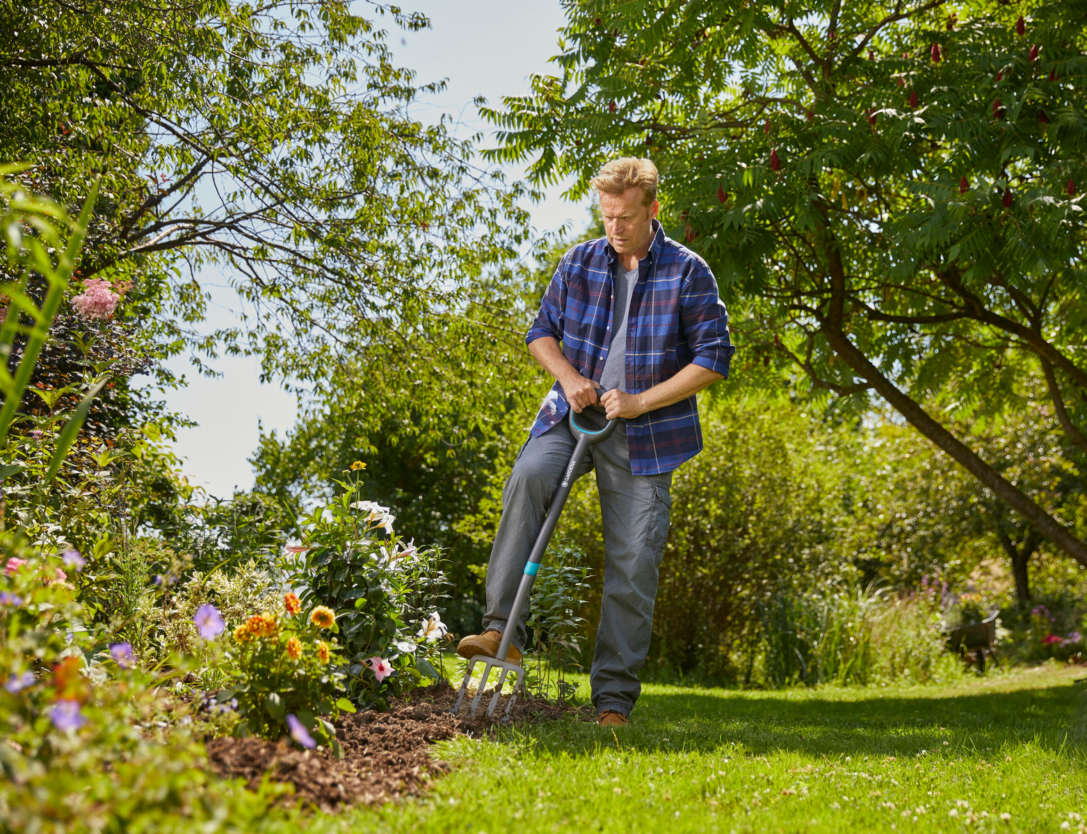 Uomo in giardino che usa una forca. Alberi e fiori sullo sfondo.