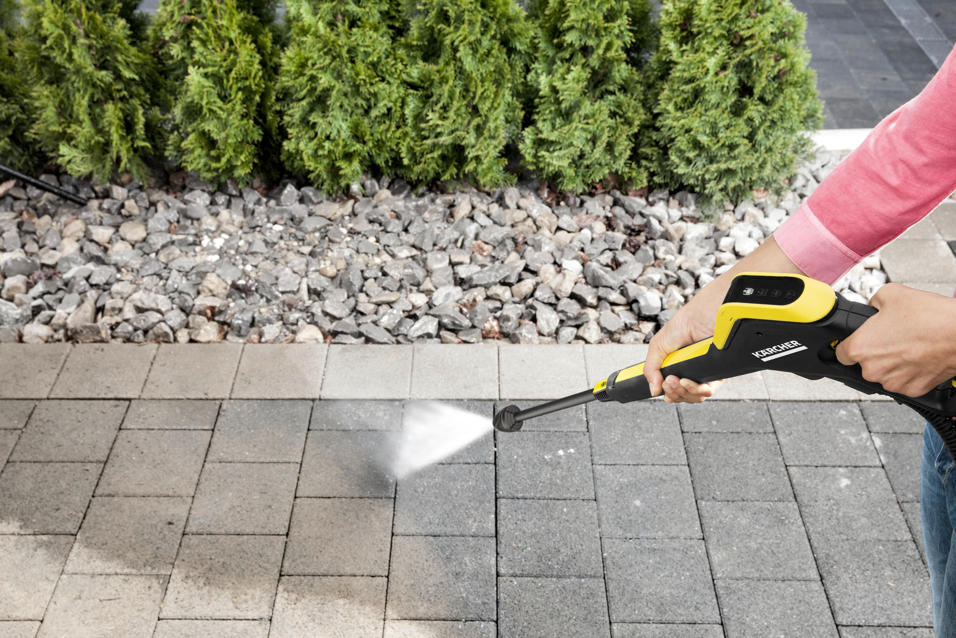 Osoba używająca żółto-czarnej myjki ciśnieniowej na szarych kostkach brukowych. Zielone rośliny w tle.