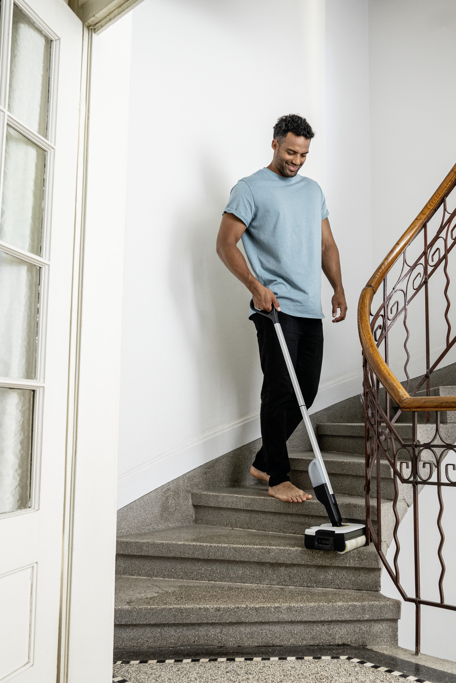 Un uomo che pulisce le scale con un aspirapolvere, a piedi nudi, sorridente, in una casa con pareti bianche e una porta.