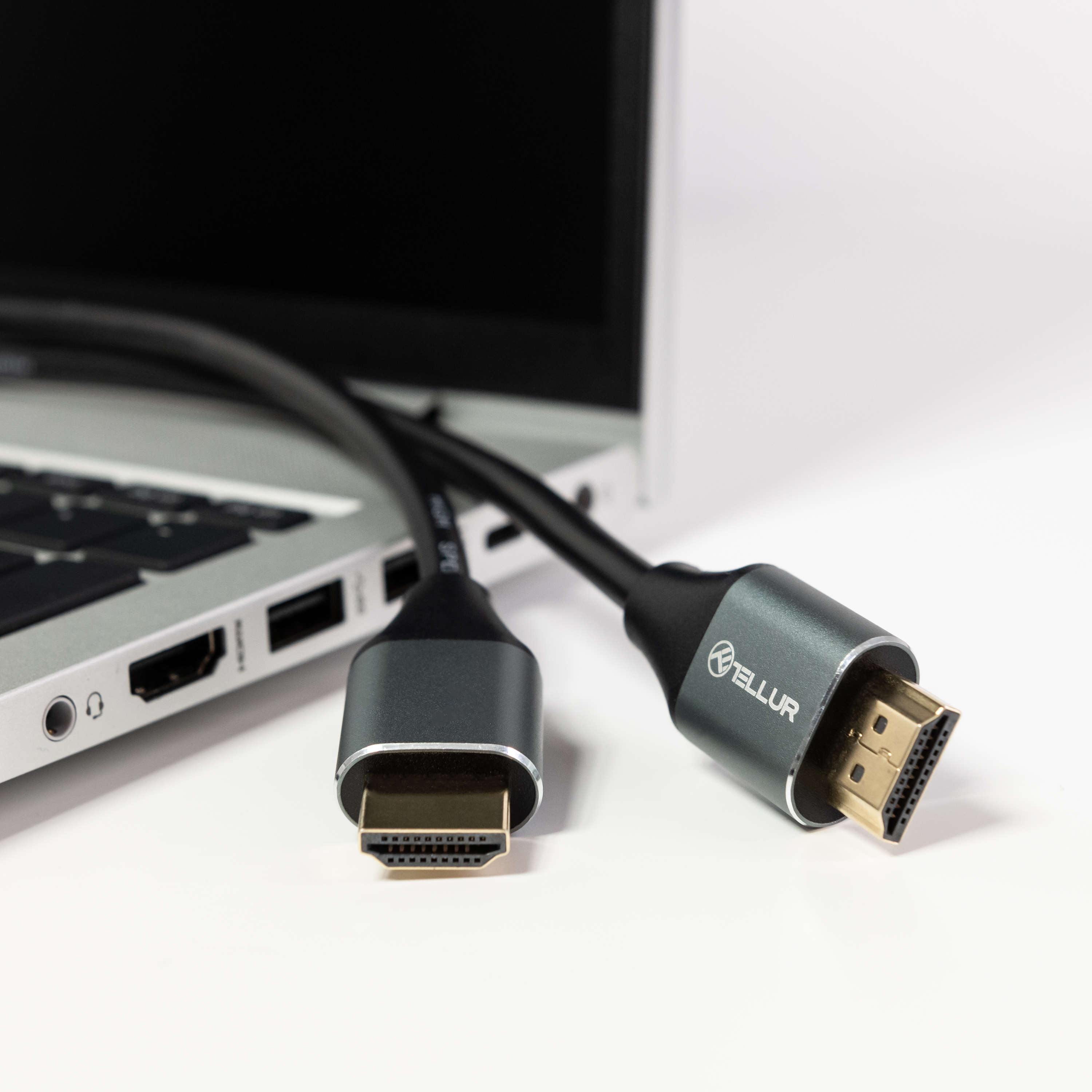 Kabel HDMI podłączony do laptopa. Czarny kabel z szarymi złączami na białej powierzchni.