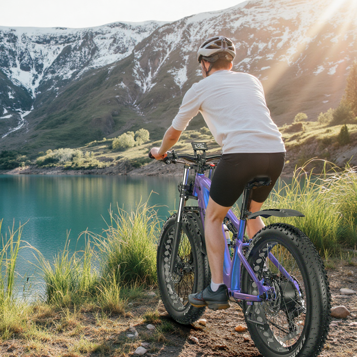 Mężczyzna na rowerze nad jeziorem. W tle góry ze śniegiem. Rower jest fioletowy, mężczyzna ma na sobie białe ubranie.