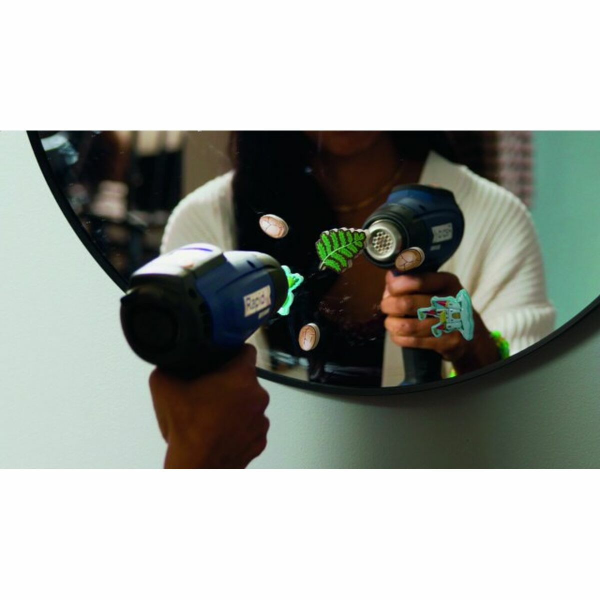 Osoba trzymająca dwa niebieskie opalarki przed lustrem. Na lustrze są naklejki.