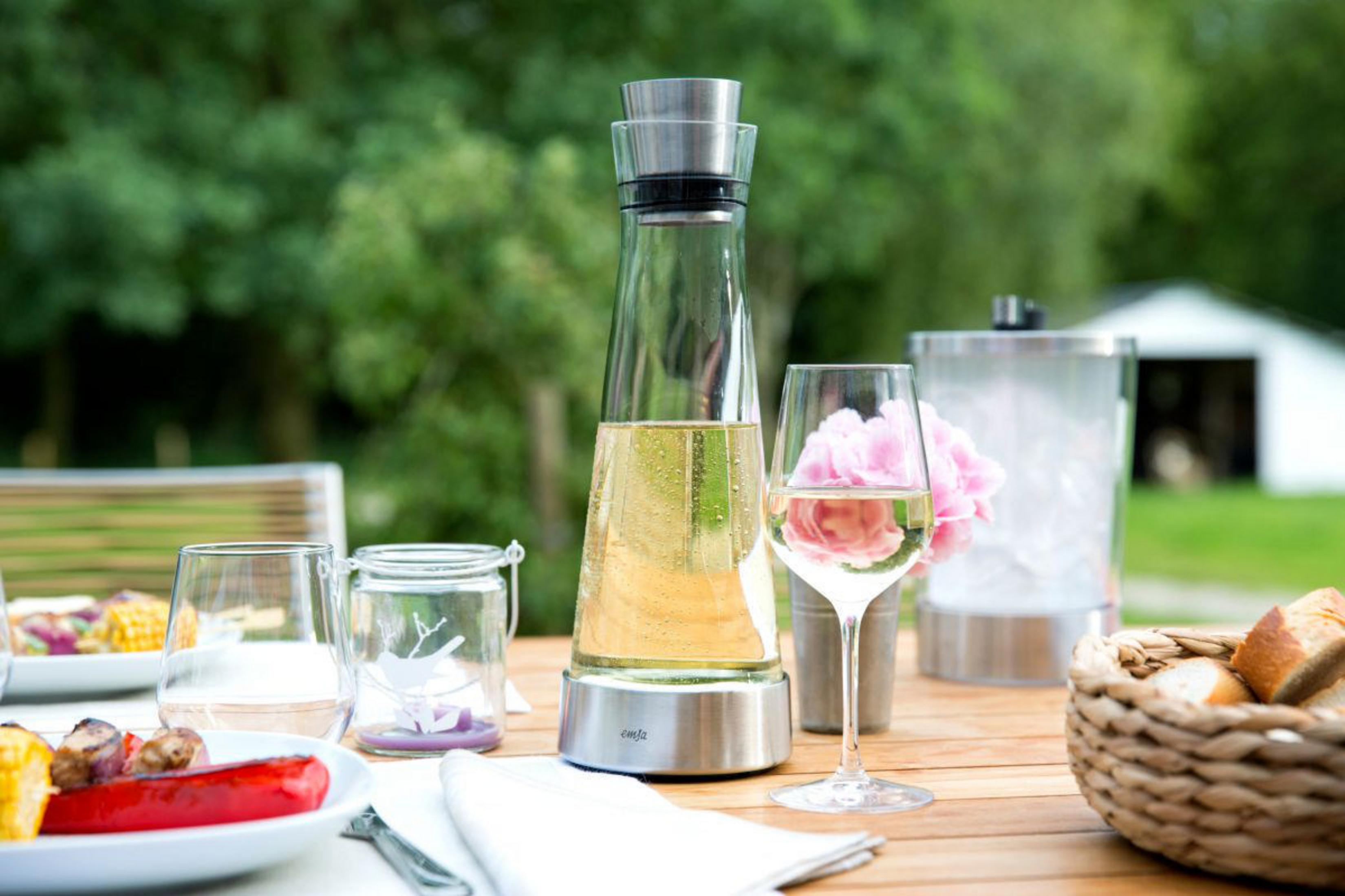 Ein Tisch mit Wein, Dekanter, Gläsern, Essen und grünem Laub im Hintergrund gedeckt.