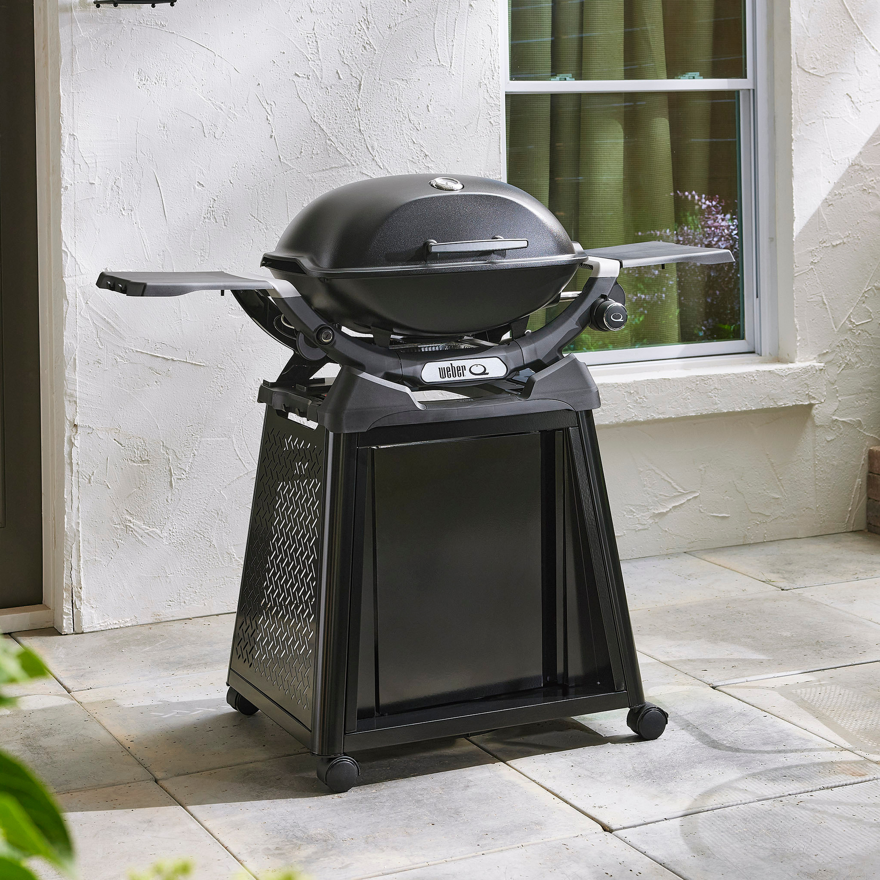 Parrilla negra Weber Q series, sobre soporte negro, al aire libre, cerca de un edificio.