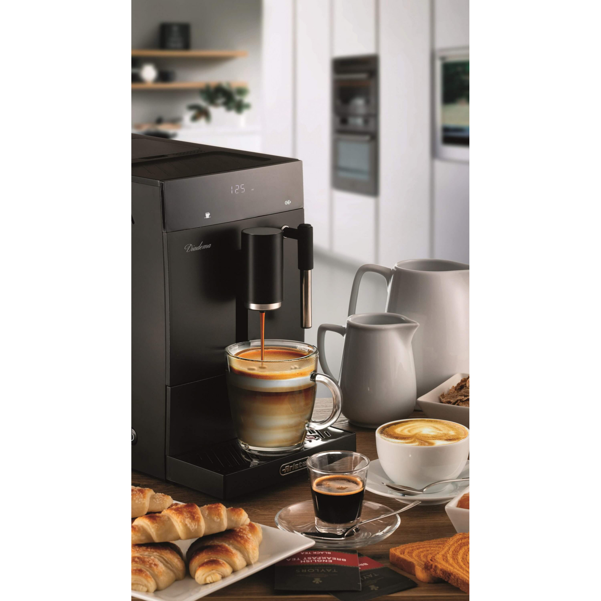 Máquina de espresso vertiendo en un vaso. Cruasanes y tazas de café en la mesa. Cocina en el fondo.