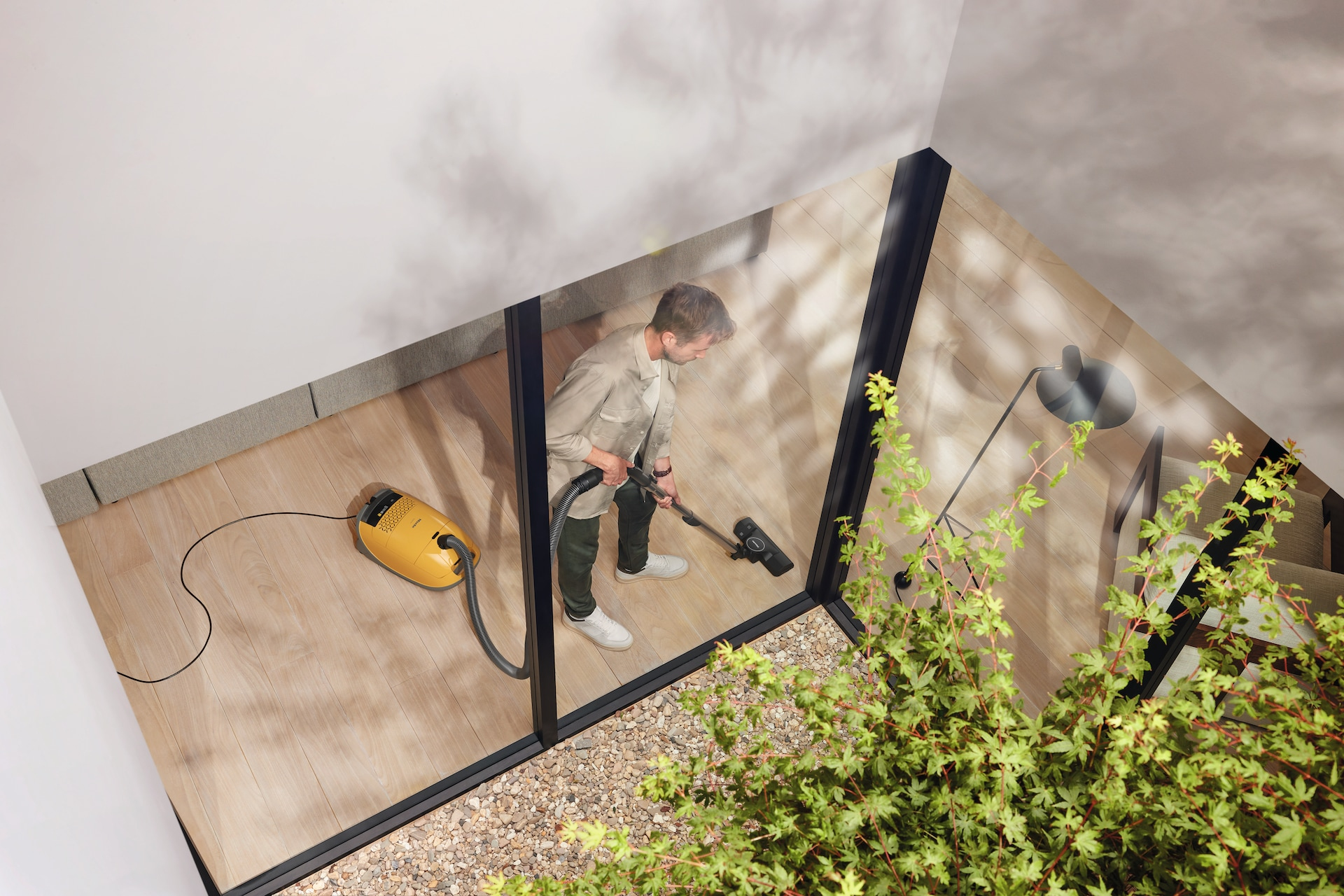 Mężczyzna odkurzający pokój żółtym odkurzaczem przez szklane drzwi.