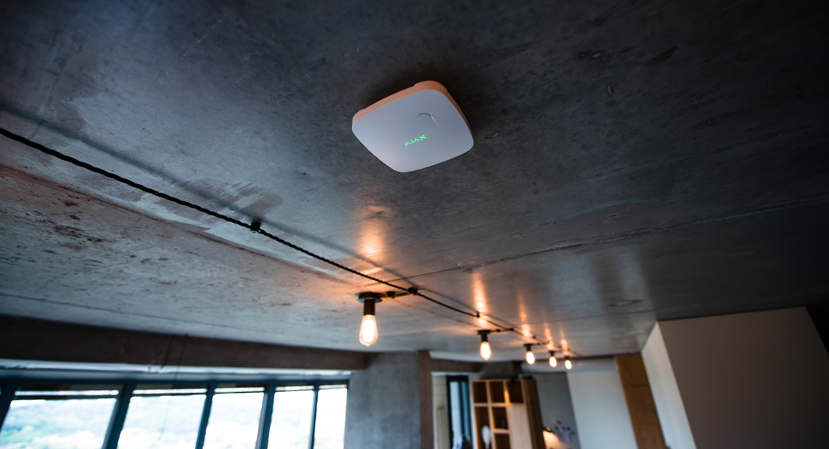 Dispositivo Ajax bianco fissato al soffitto. Fili e luci visibili sullo sfondo.